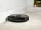 A black robotic vacuum cleaner is cleaning the beige carpet along a white wall, sucking up small crumbs.