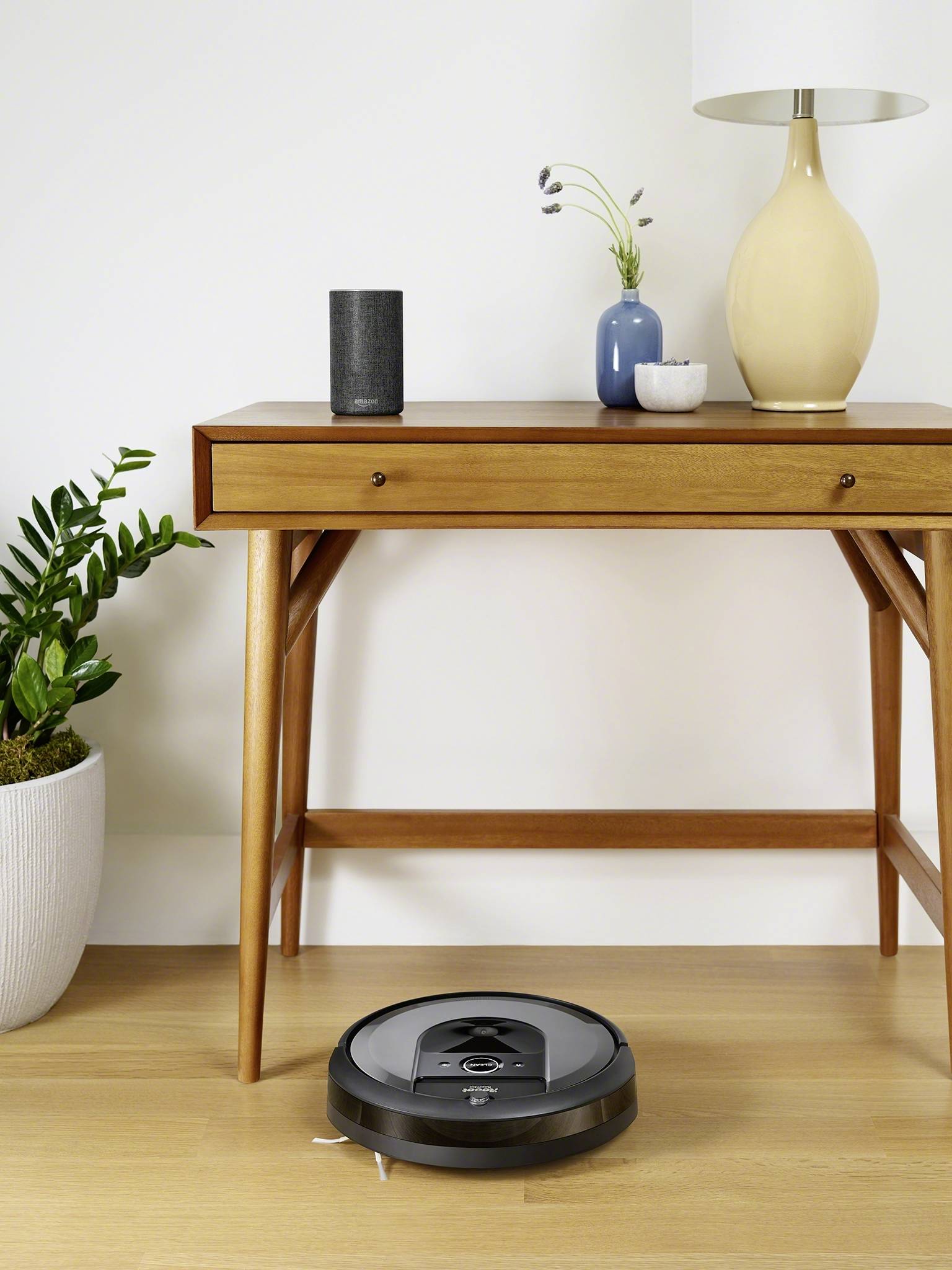 A robot vacuum cleaner is moving across a wooden floor in front of a table with a lamp and plants. A plant pot is standing on the left.