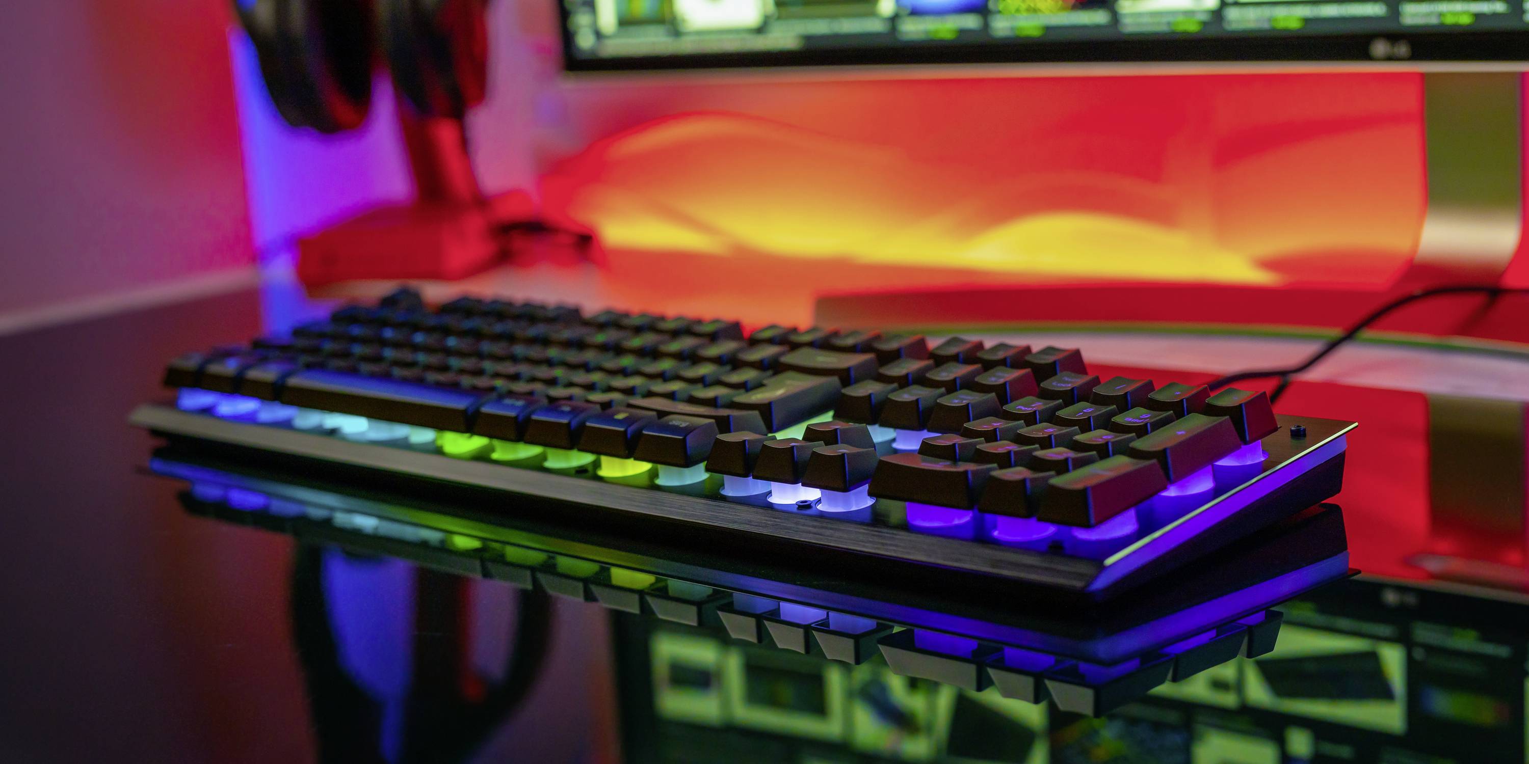 A backlit mechanical keyboard with RGB lights on a desk in front of a large computer monitor in a colourful room.