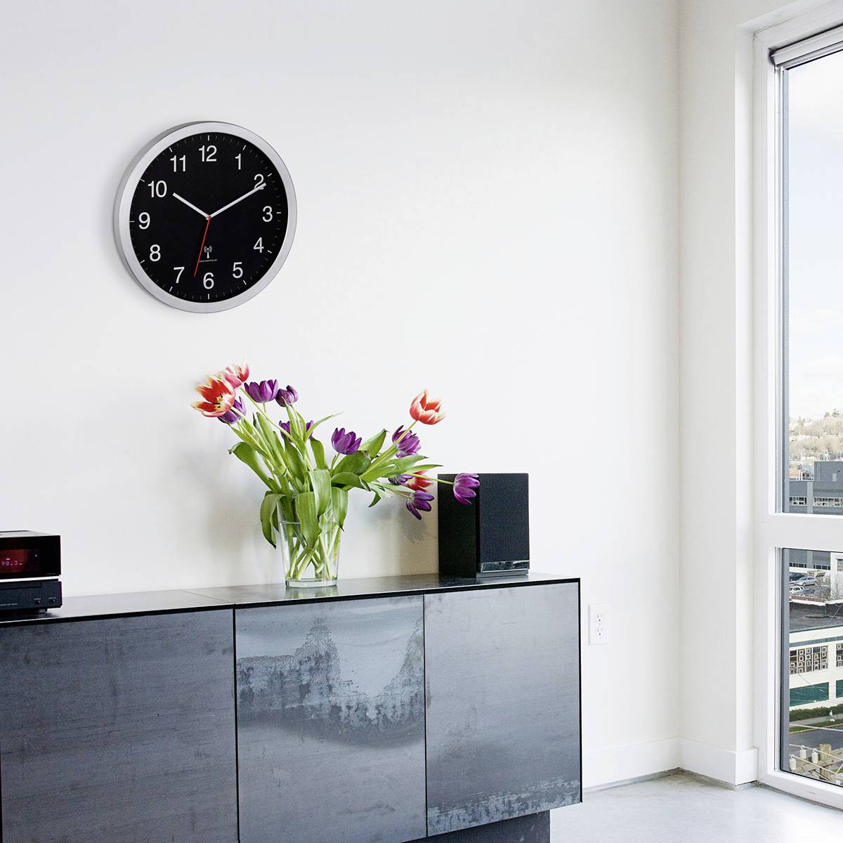 White room with a large clock on the wall, showing 10:10. A sideboard underneath with a flower bouquet and camera, window on the right with a city view.