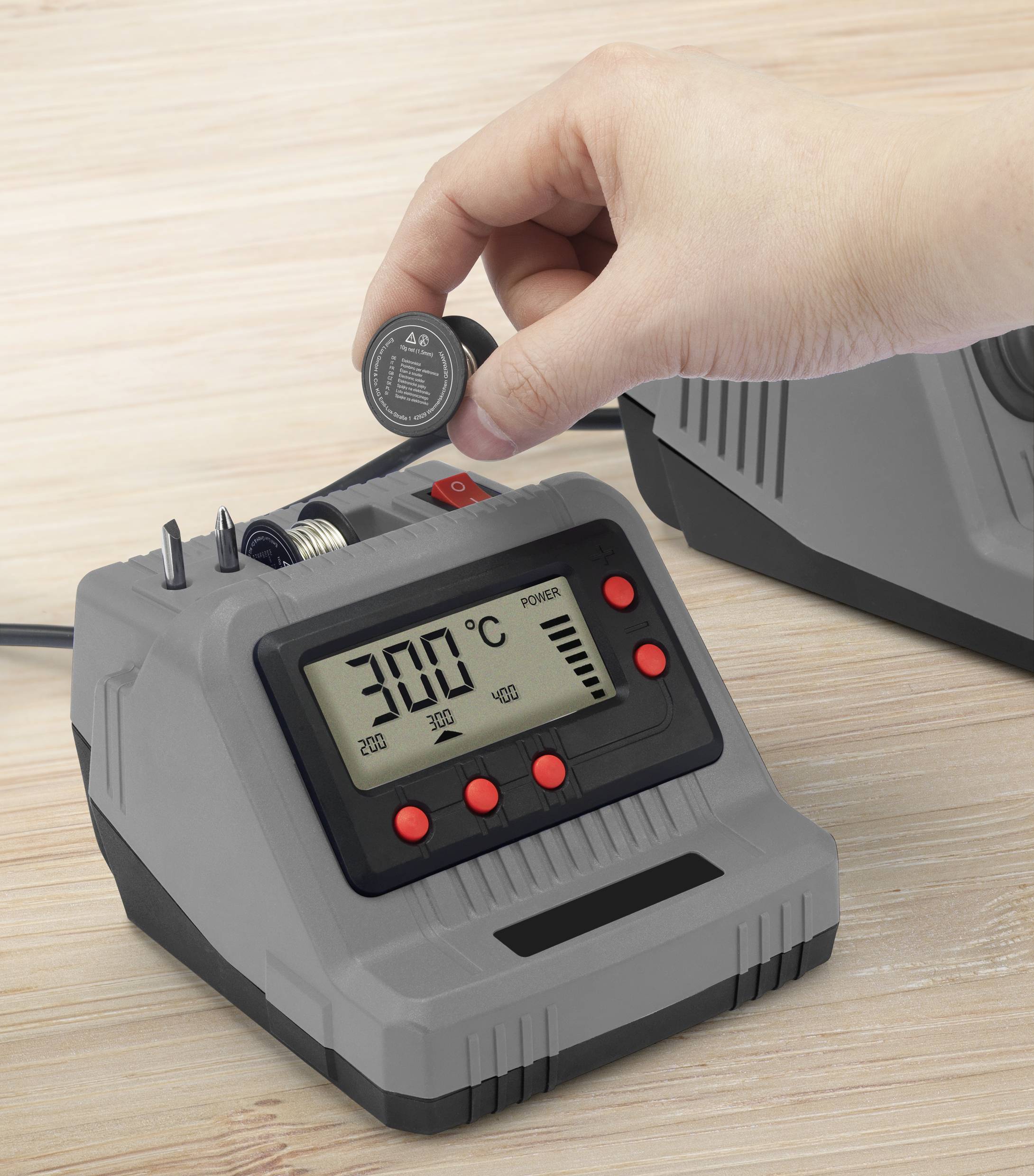 A person is holding a battery over a digital charger on a wooden table. The display shows '300°C' and charging information.