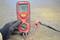 A red multimeter displays '0.07'. A gloved hand is holding the device, which is connected to two cables with clamps.
