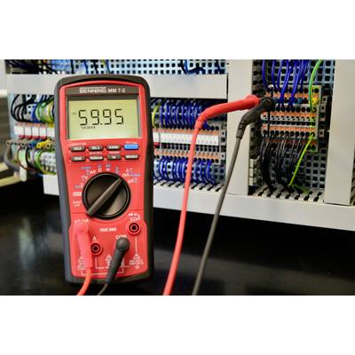 Multimeter on a table displays '-59.95'. In the background, an electrical cabinet with cables and connections. Device for measuring electrical values.