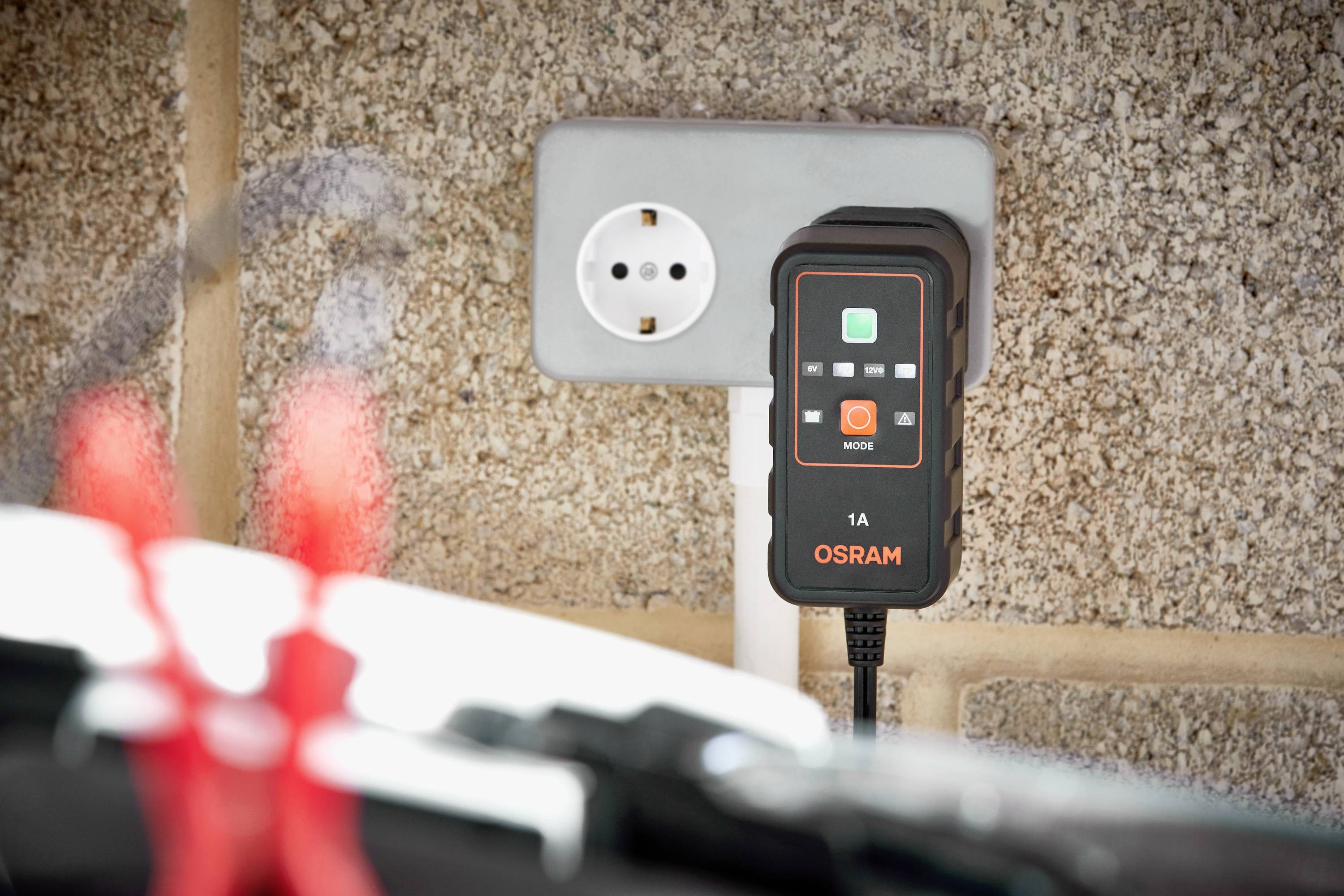 An Osram battery charger is plugged into a wall socket. In the foreground, blurry red clips are visible.