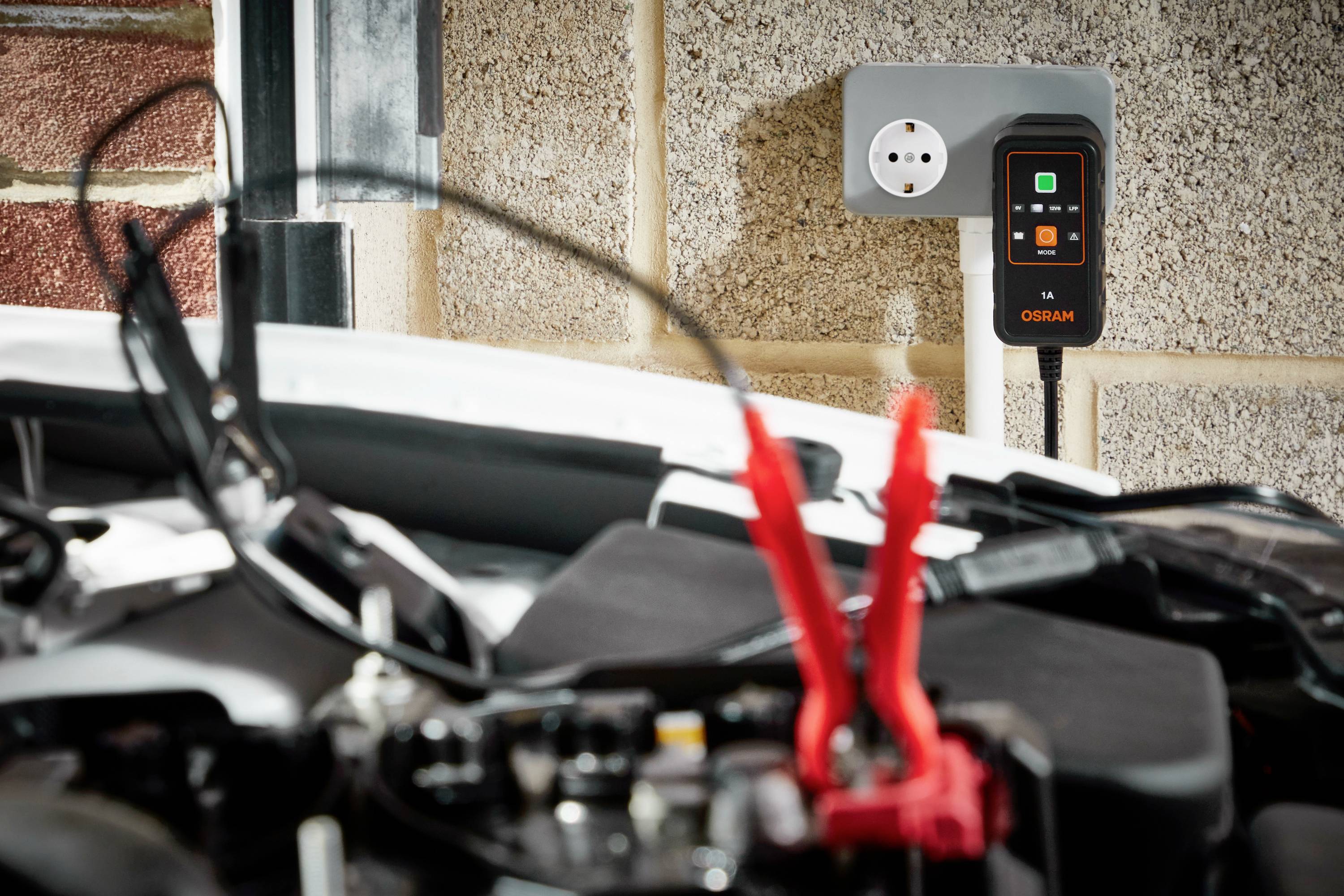 A charger is plugged into a socket, while clamps are attached to a car battery, possibly for charging.