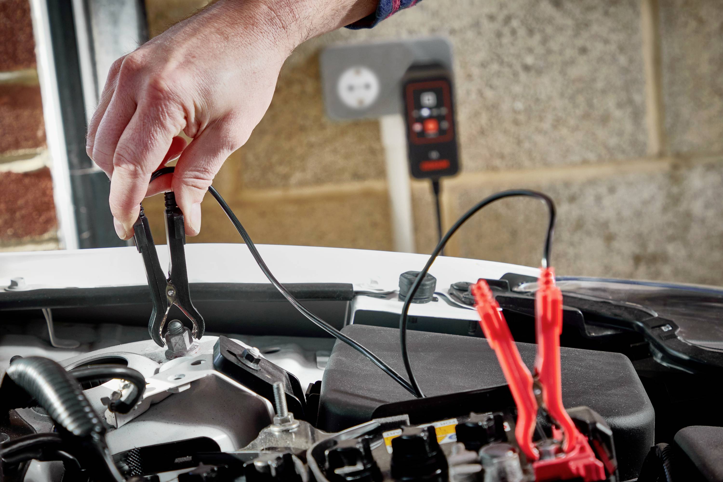 A hand is holding a jump lead clip on a car battery, with a second clip already connected. A charger is visible in the background.