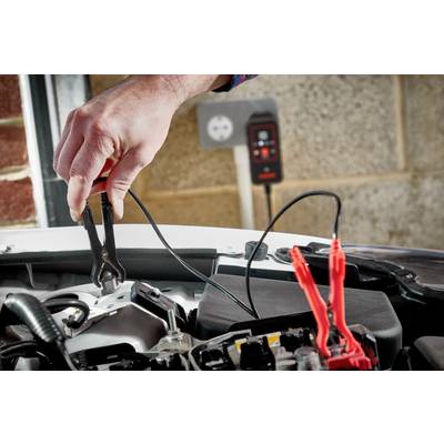 A hand is holding a jump lead clip on a car battery, with a second clip already connected. A charger is visible in the background.