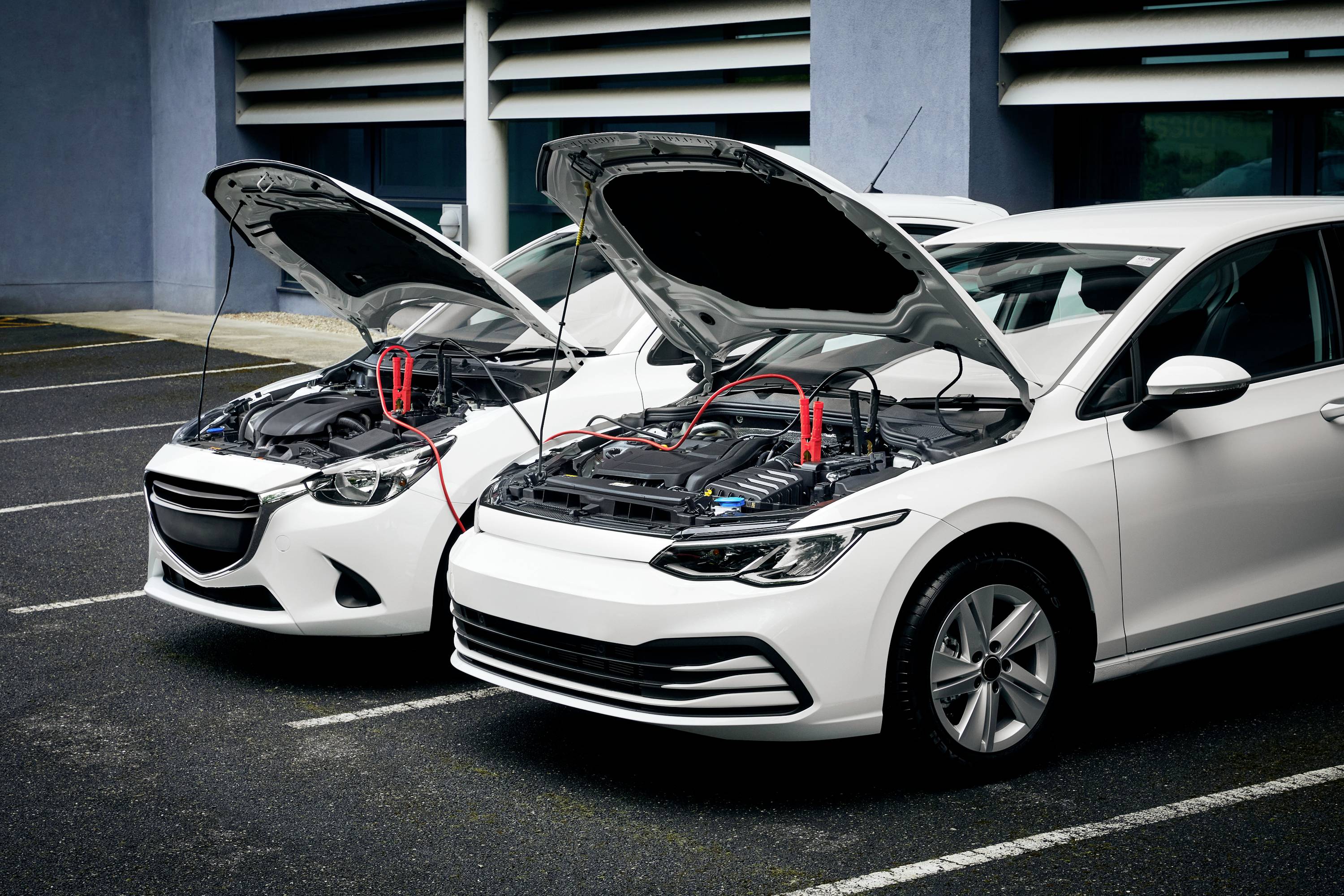 A person is starting a car by jump-starting it with another vehicle. Both bonnets are open and the cables are connected.