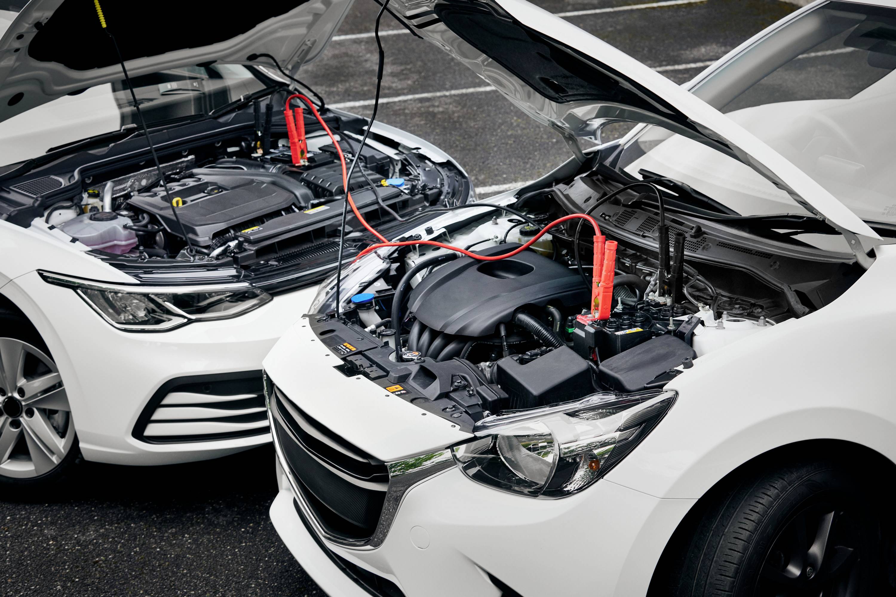 Two cars with open bonnets, connected by jump leads. The car on the left is giving a jump start to the car on the right.