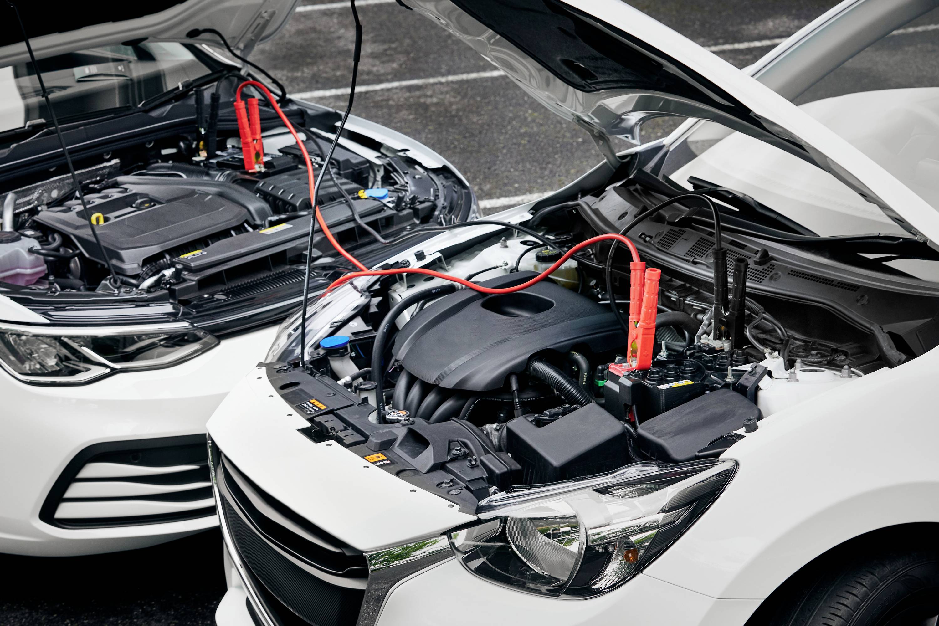 Two cars are parked side by side with open bonnets, connected by red jump leads.