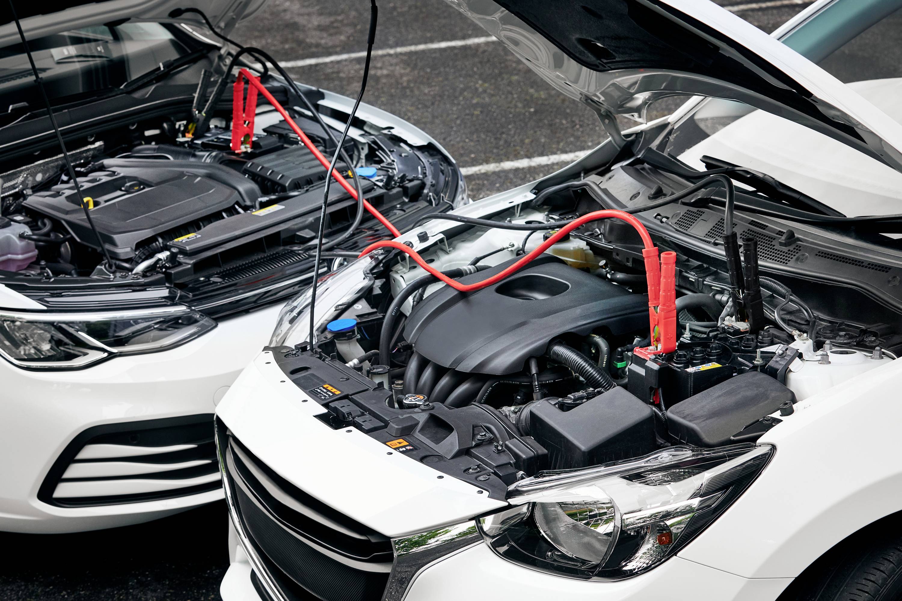 Two cars with open bonnets in a car park. Jump leads connect the batteries to start one vehicle.