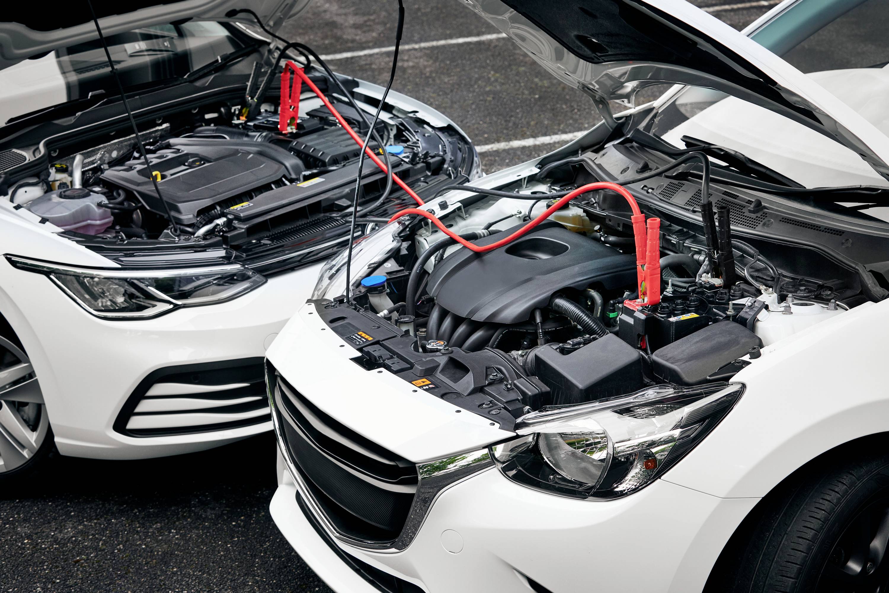 Two cars are parked side by side with their bonnets open. Jump leads are connected to charge a battery.