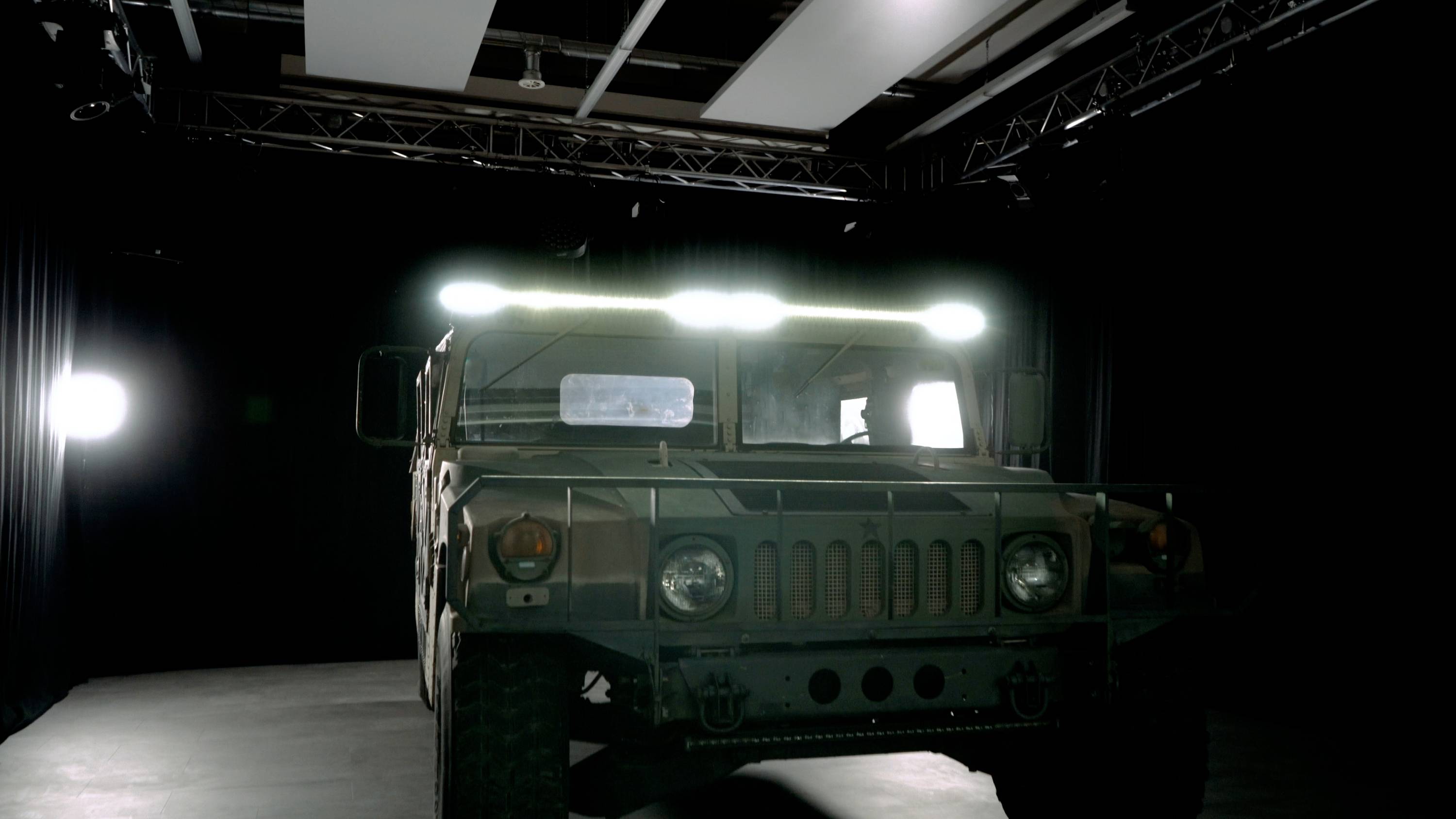 An armoured military vehicle stands in a dimly lit room, surrounded by spotlights. The light accentuates its robust construction.
