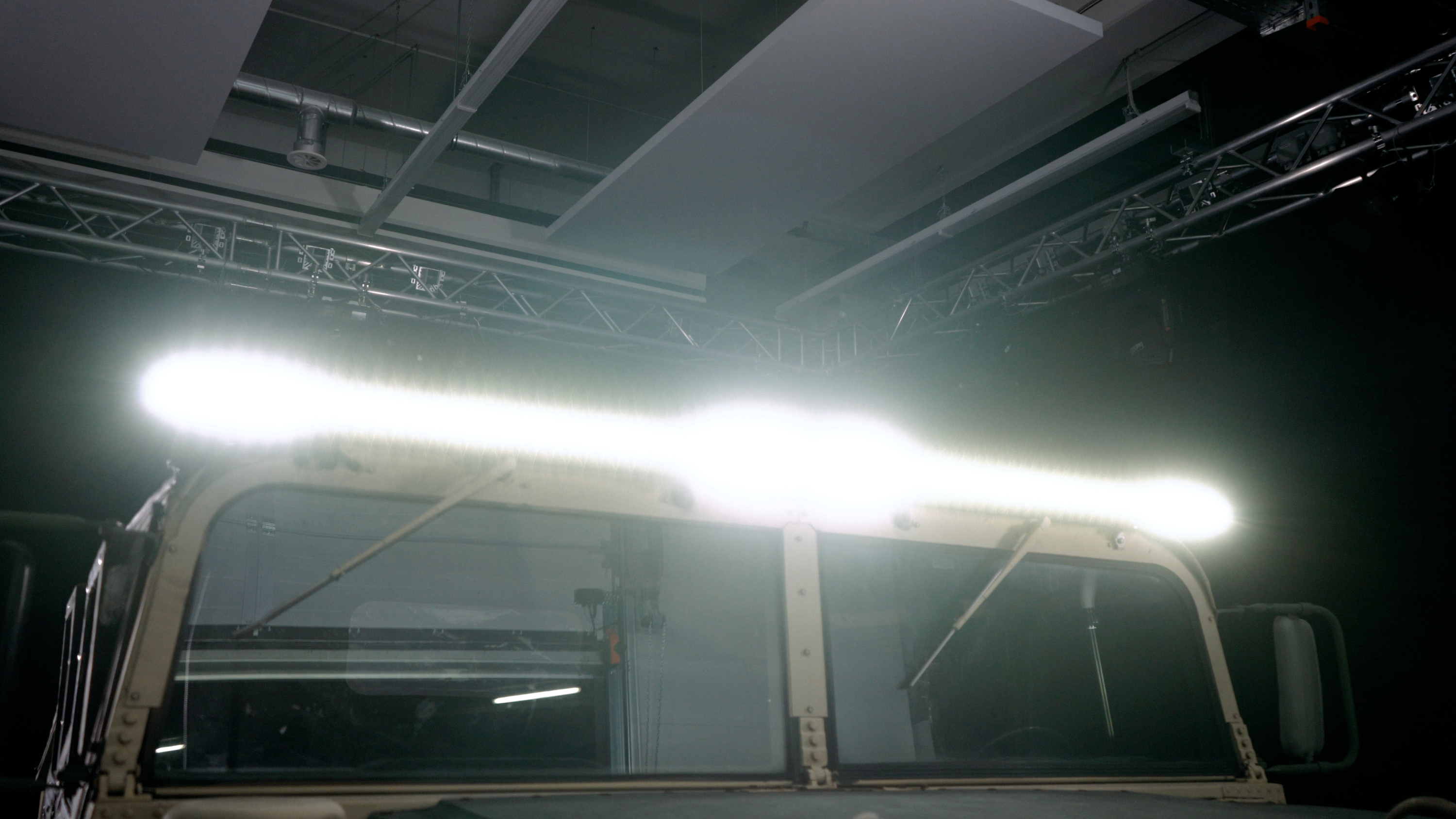 An off-road vehicle with a brightly illuminated LED light bar on the roof, stands in an industrial space with visible pipework and lighting.