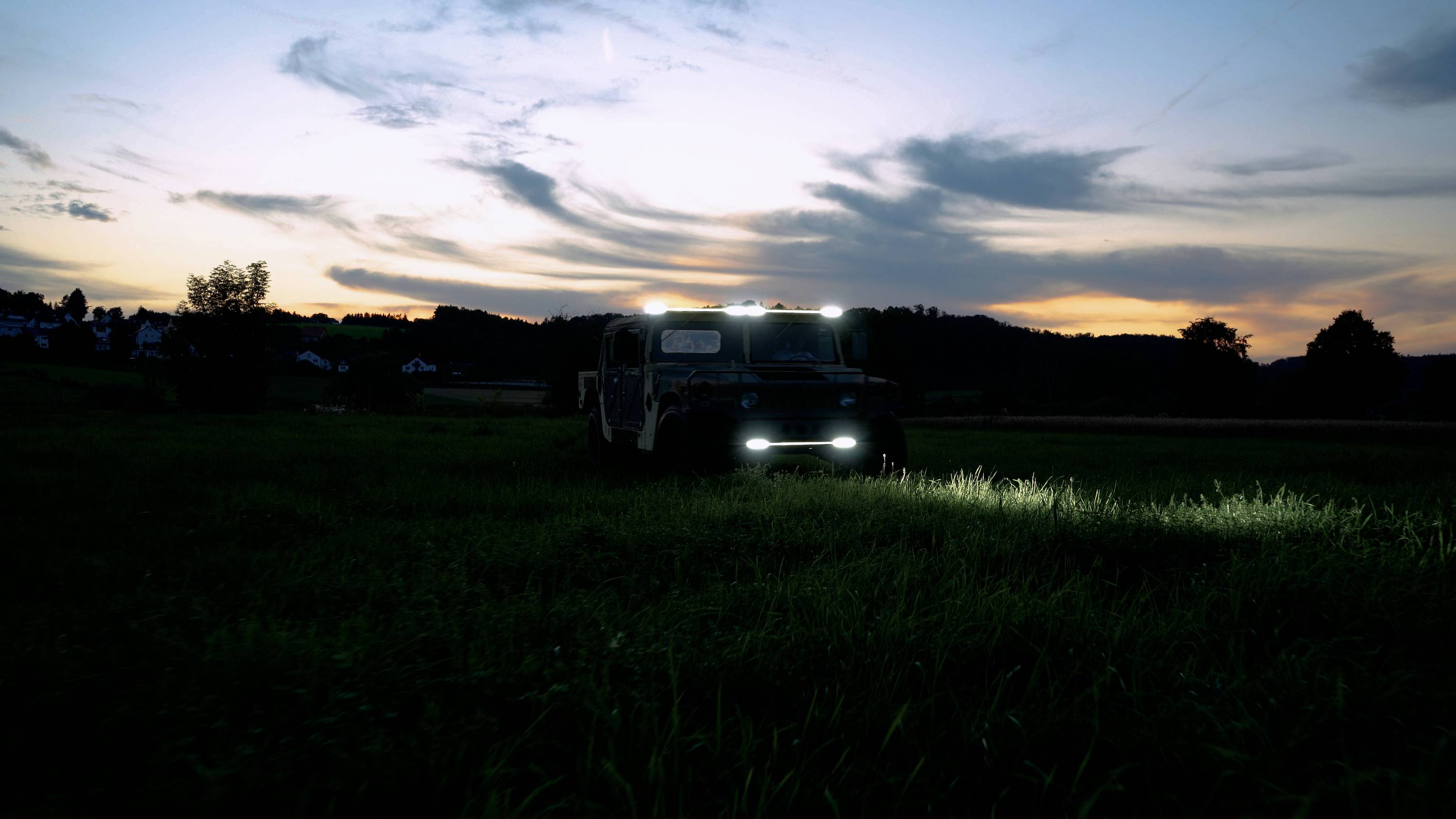 A vehicle with its lights on stands on a meadow at dusk. The sky displays a colourful sunset.