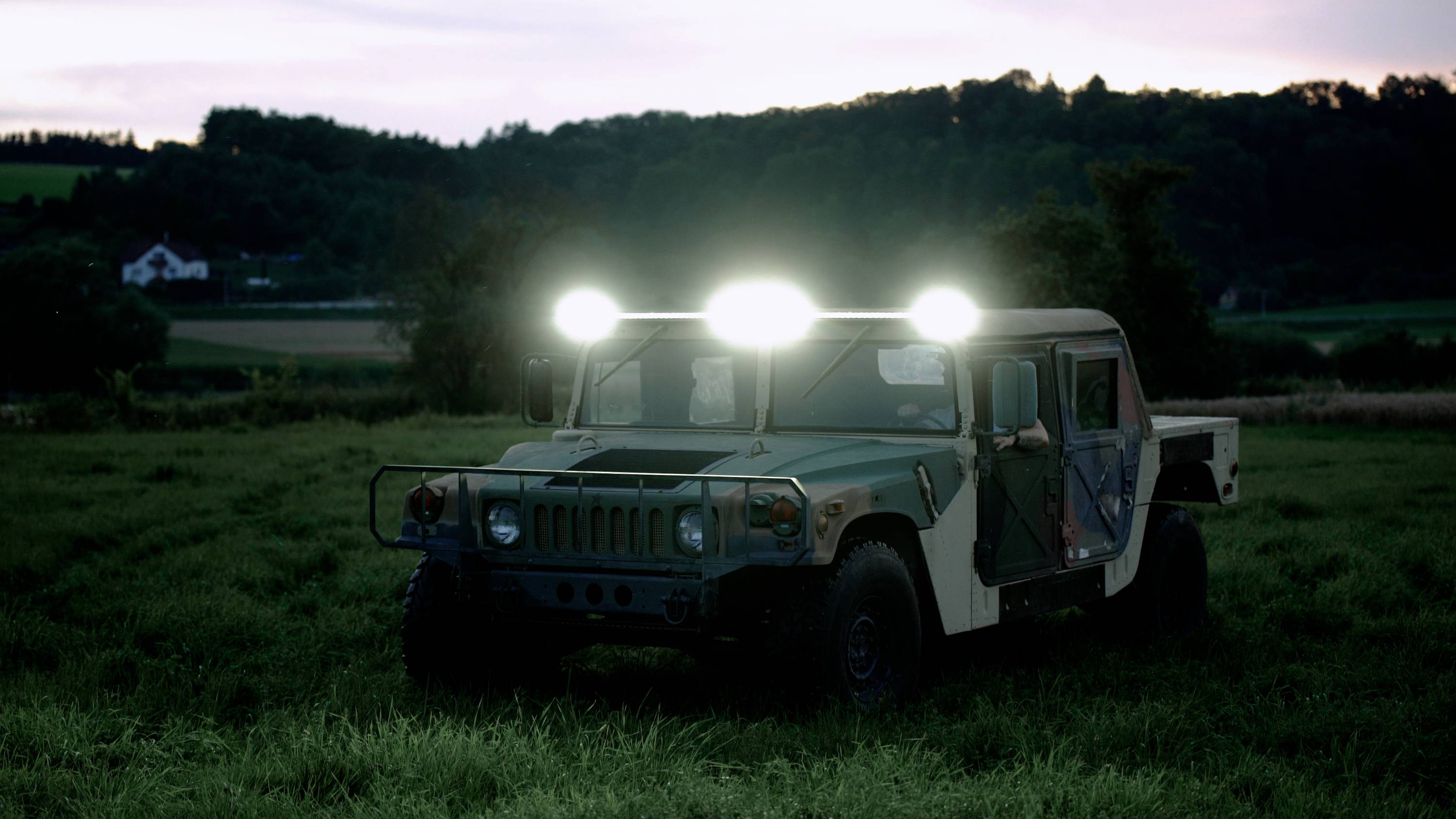 A military vehicle with its headlights on stands on a meadow at dusk, surrounded by trees and hills.