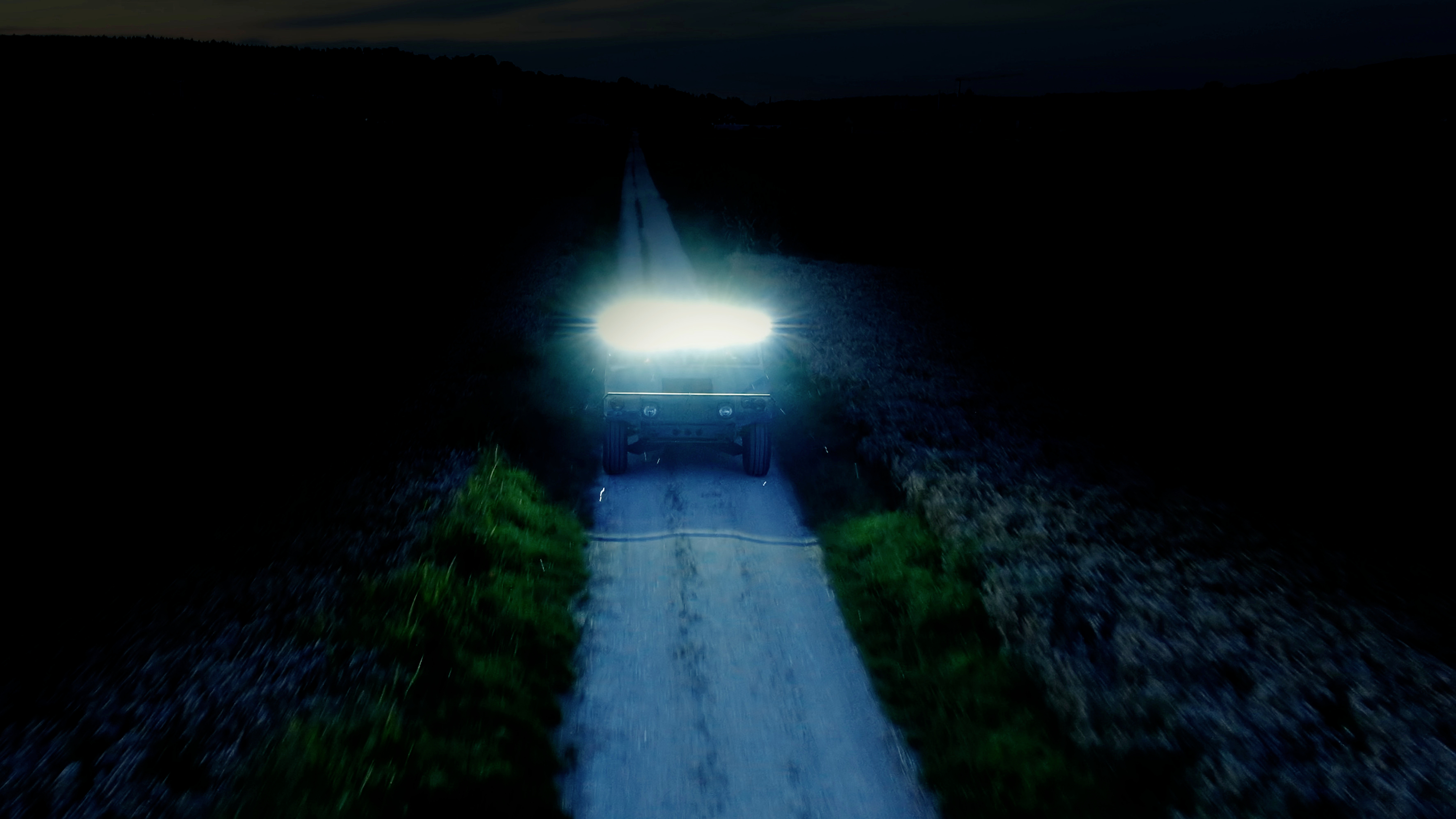 A vehicle with bright headlight beams drives along a deserted road at night. The surroundings are dark and unlit.