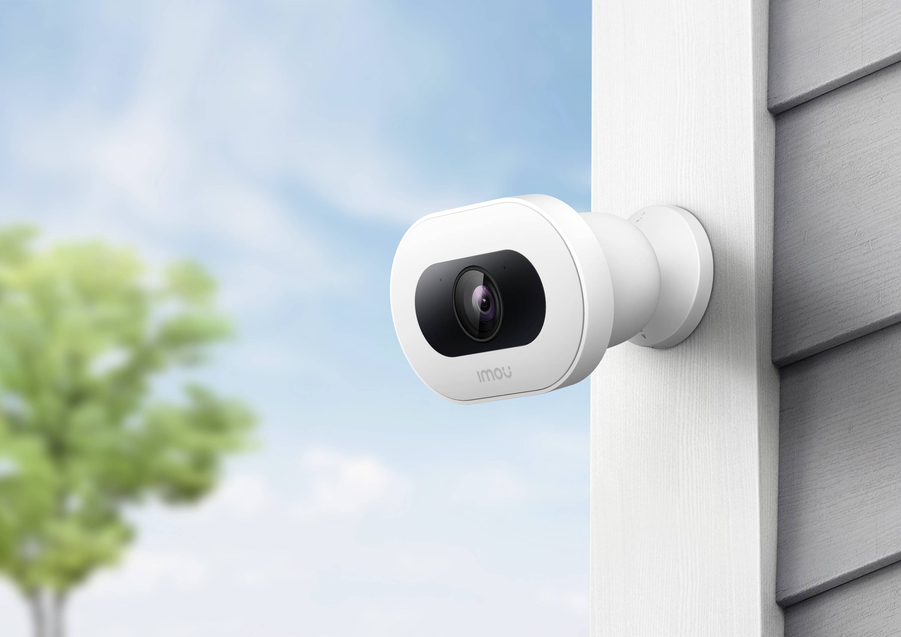Security camera mounted on a house wall, with a blurry tree and blue sky in the background.