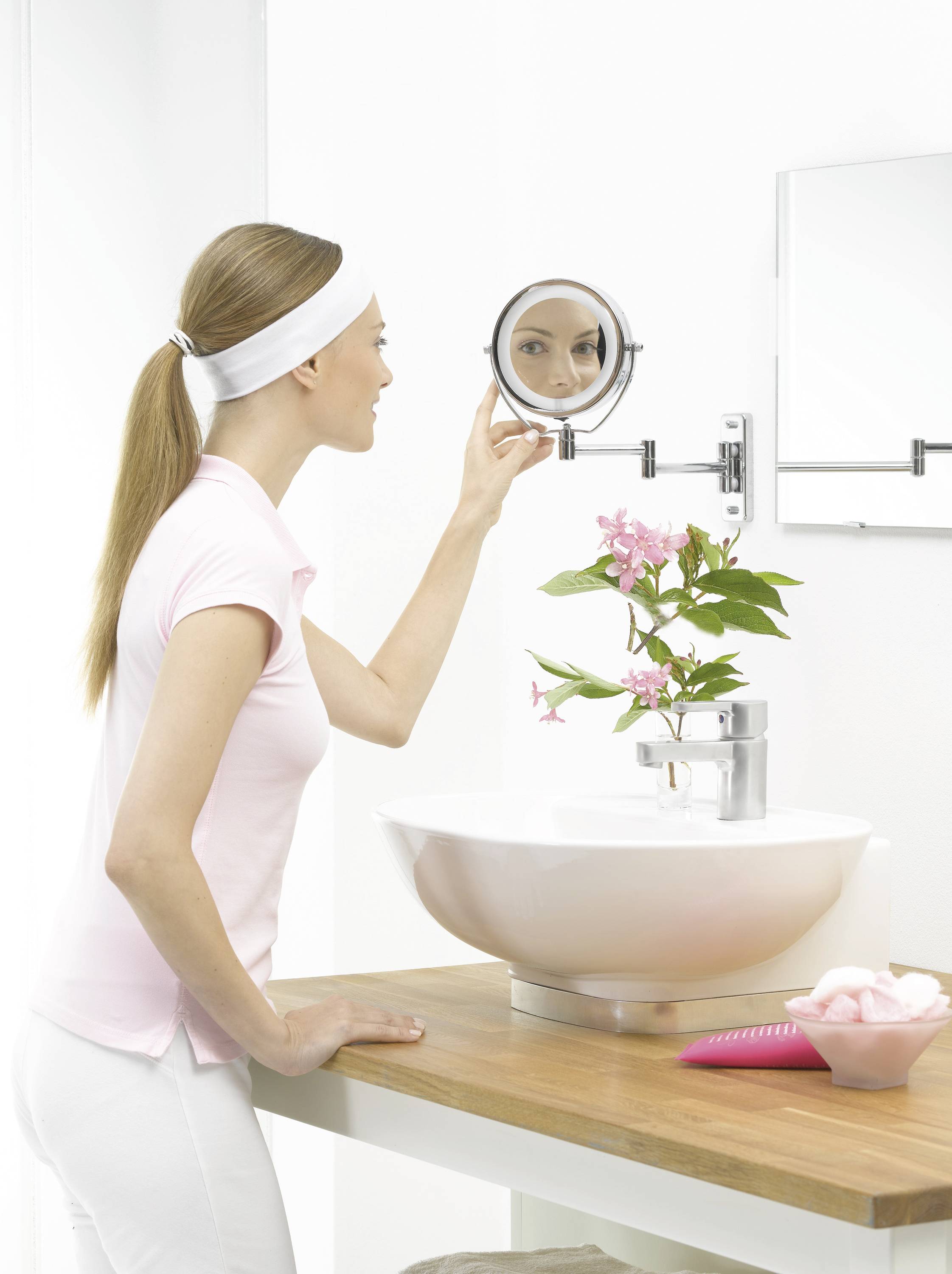 A woman looks at herself in a round wall mirror in the bathroom, surrounded by white walls and a flower in a vase next to the sink.