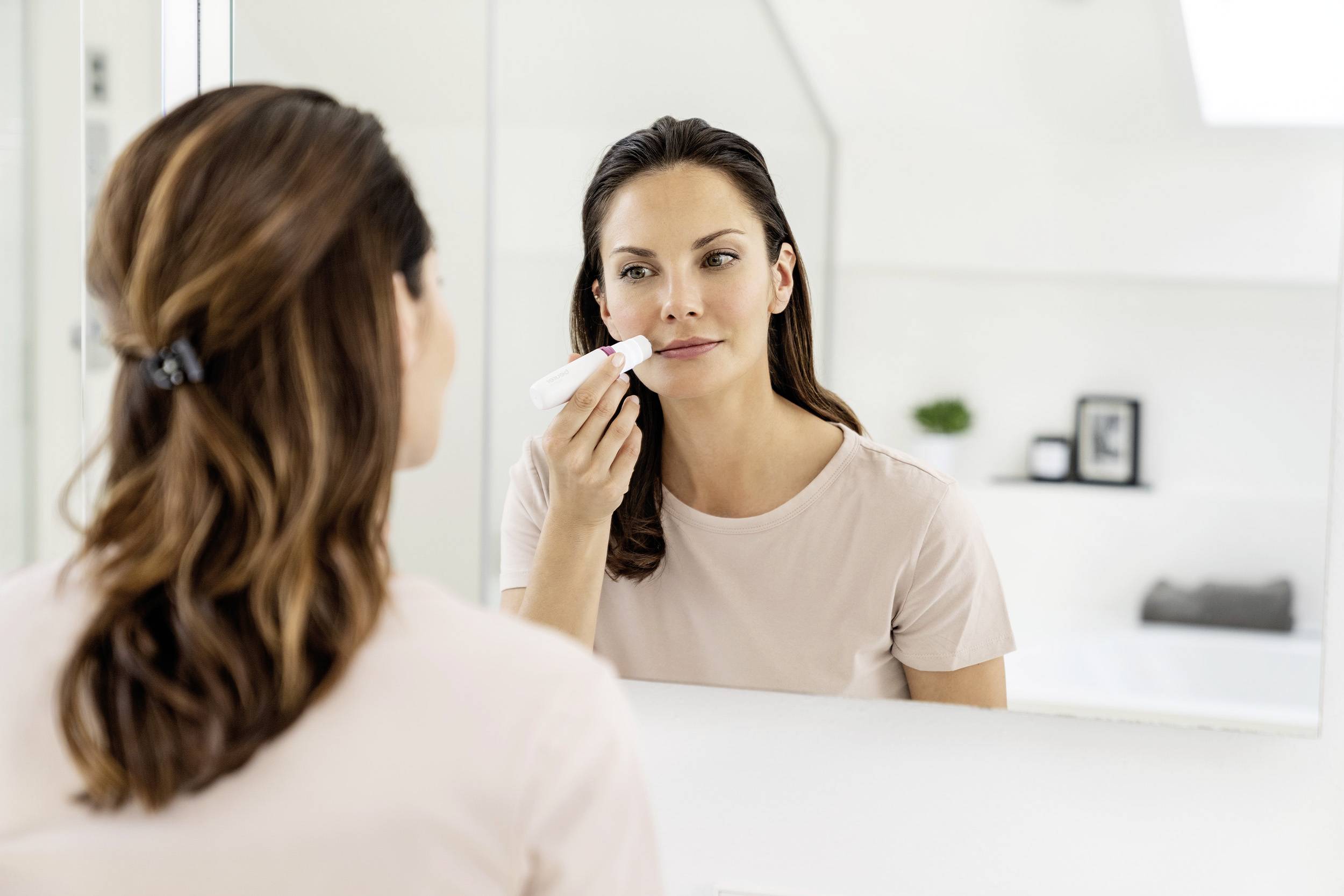 A woman is using a facial massage device in front of a mirror in the bathroom. She is wearing a light-coloured top and has brunette hair.