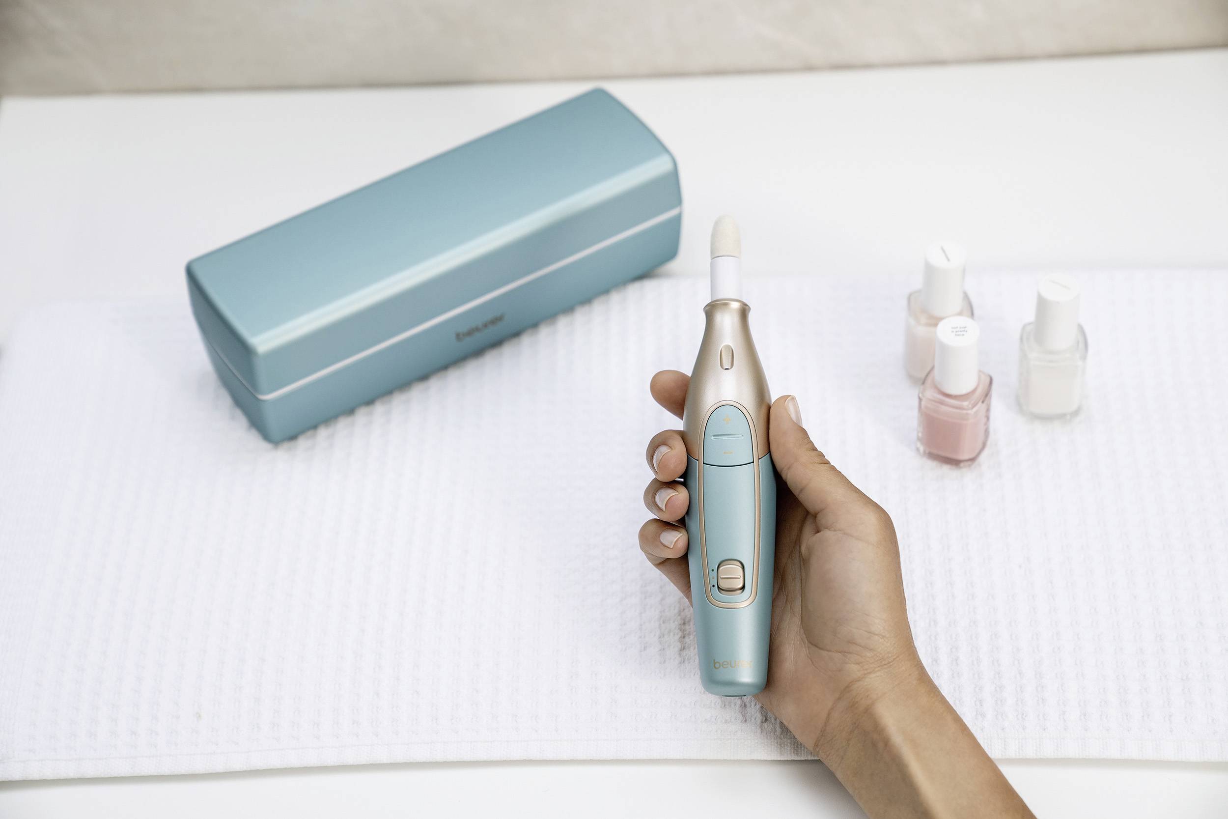 A hand is holding an electric nail care device next to painted nails and three nail polish bottles, with a case in the background.