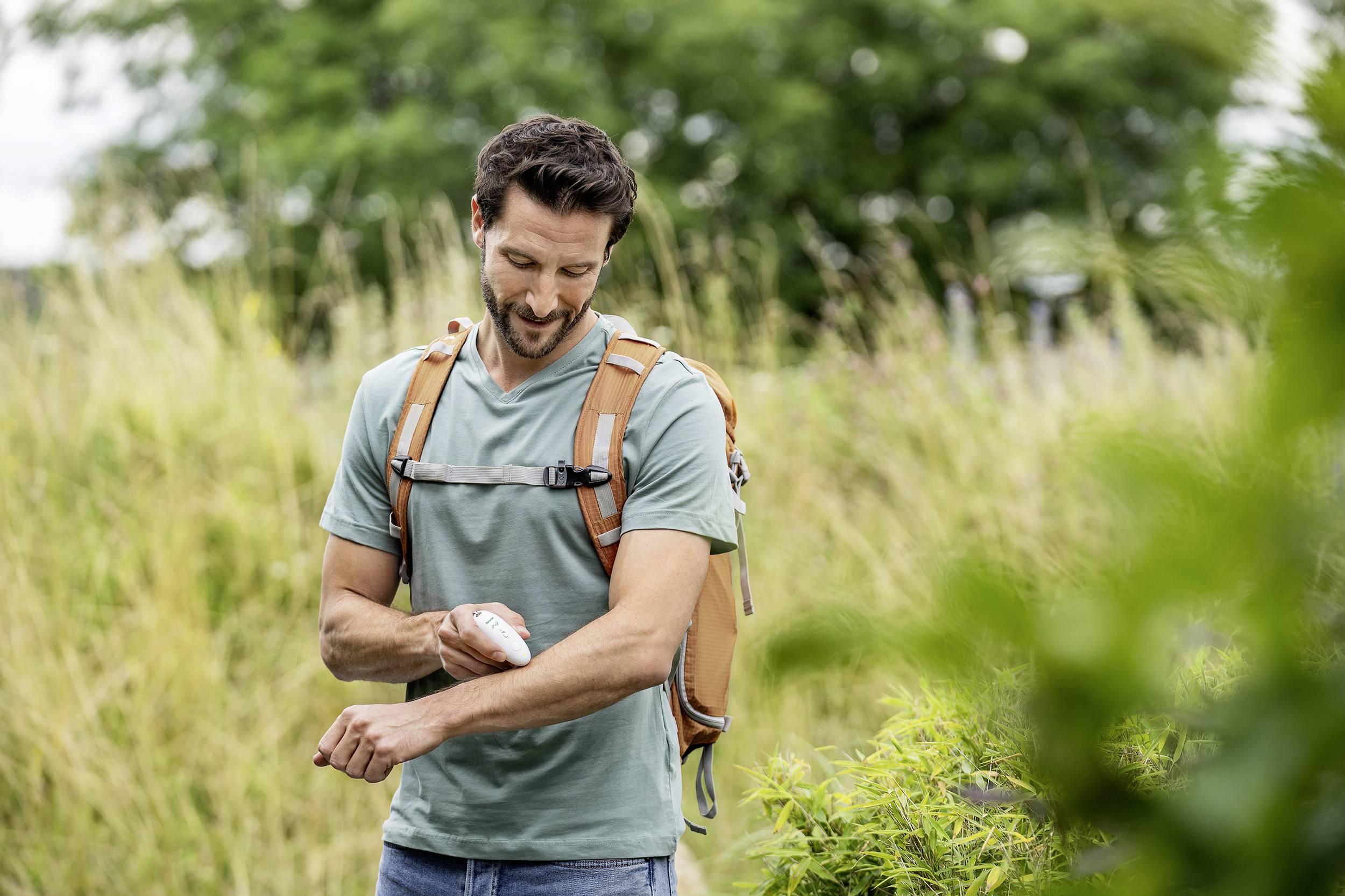 A smiling man outdoors is wearing a rucksack and applying sun cream to his arm to protect himself from the sun.