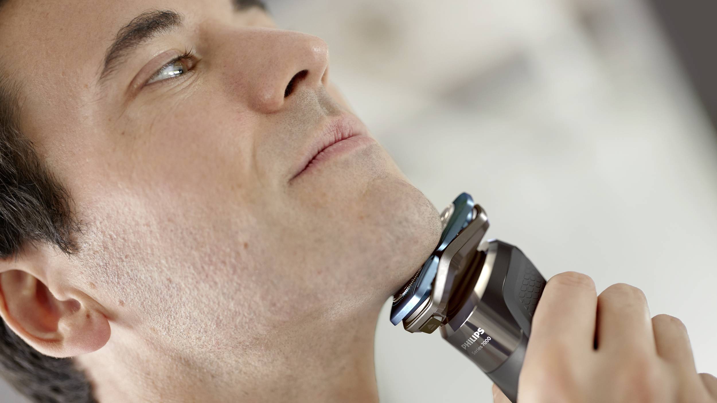 A man is shaving his chin with an electric razor. The background is out of focus.