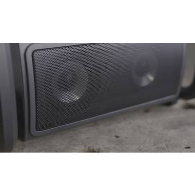 Close-up of a black, portable speaker with a metal grille. The speaker is standing on a concrete floor.