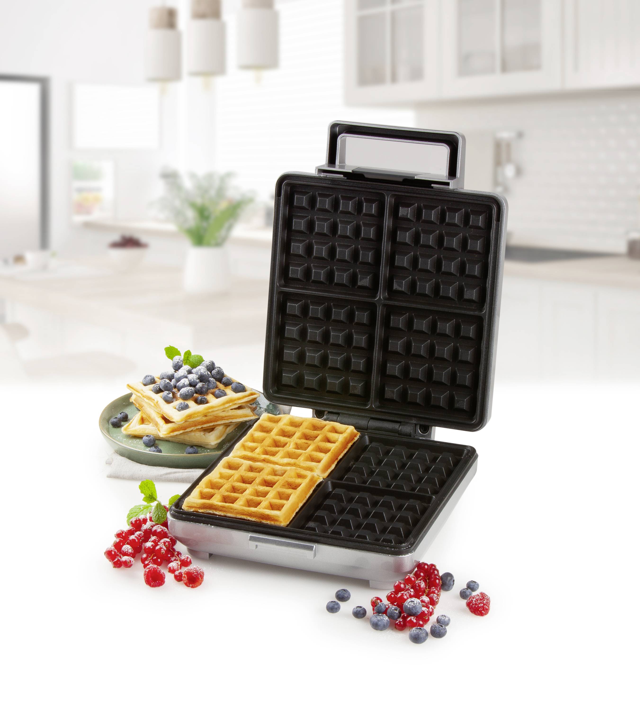An open waffle iron with two golden-brown waffles. In the background, waffles are placed on a plate, garnished with berries.