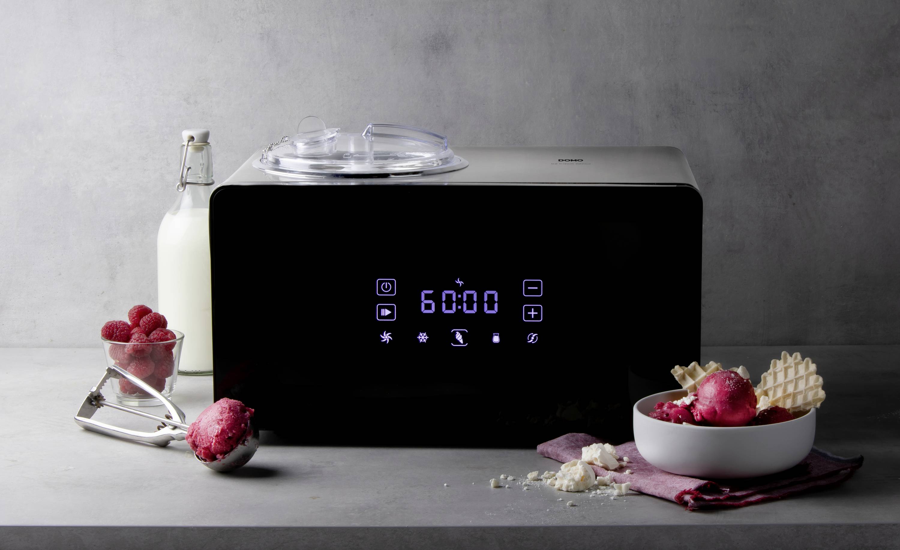 An ice cream machine with a digital display shows '60:00'. Beside it are a bottle of milk, fresh raspberries, and a bowl of ice cream.