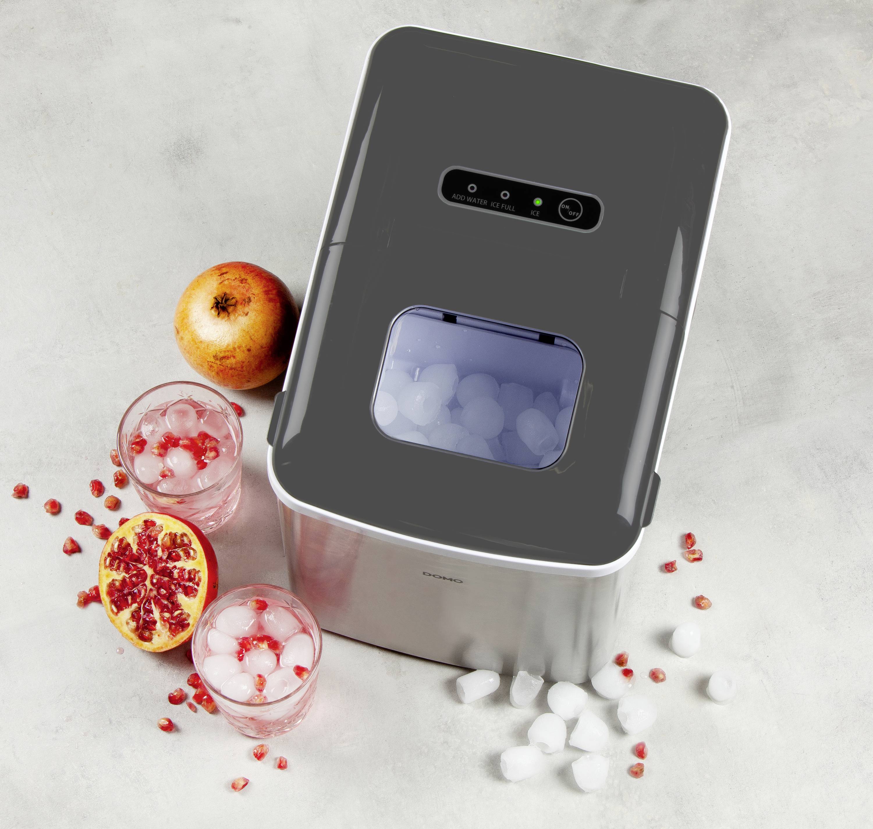 A silver, rectangular ice maker on a grey surface, surrounded by two glasses of pomegranate juice and half a pomegranate.