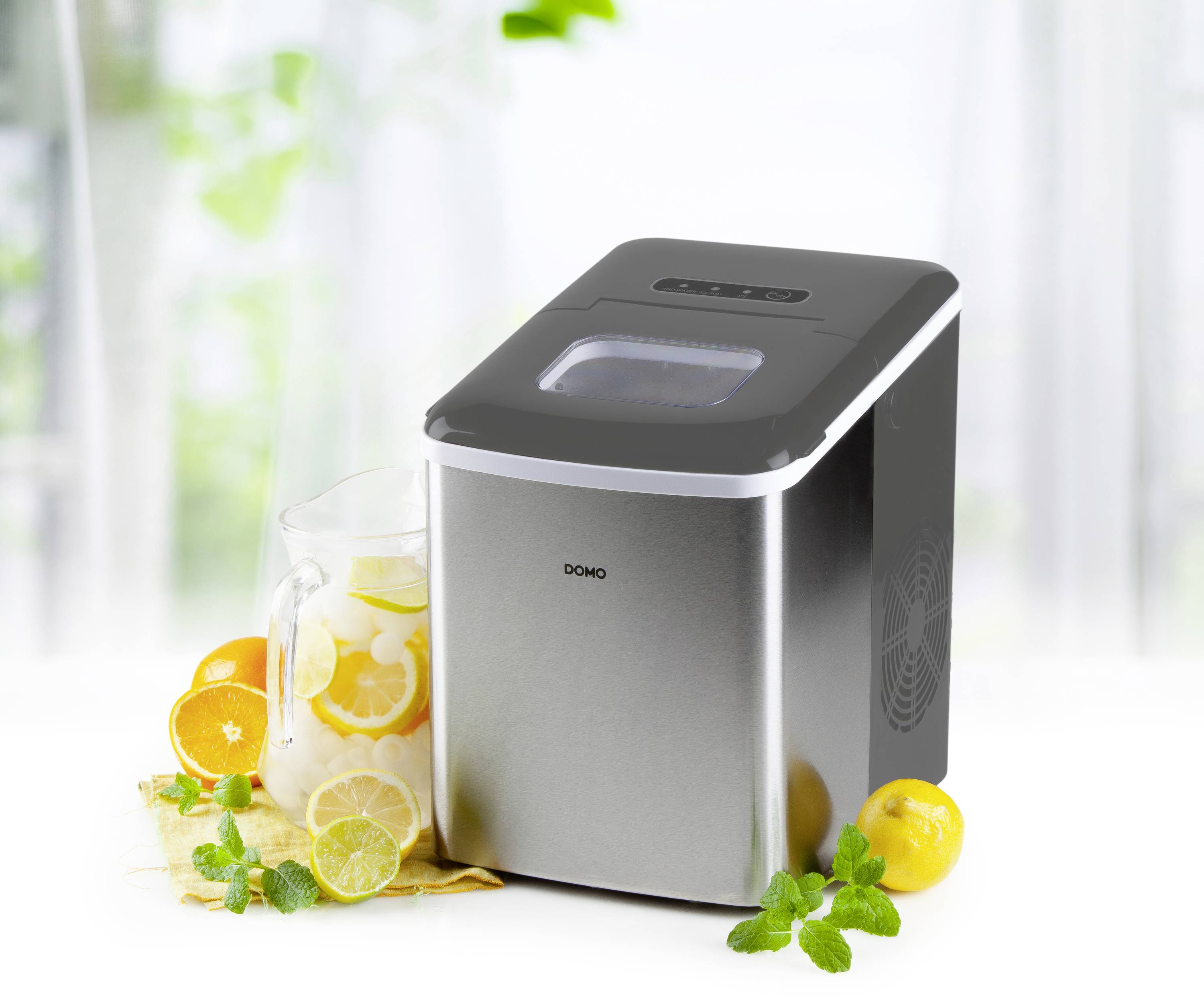 Ice cube machine next to a carafe with citrus fruits and mint. Device is placed on a white surface against a blurred background.