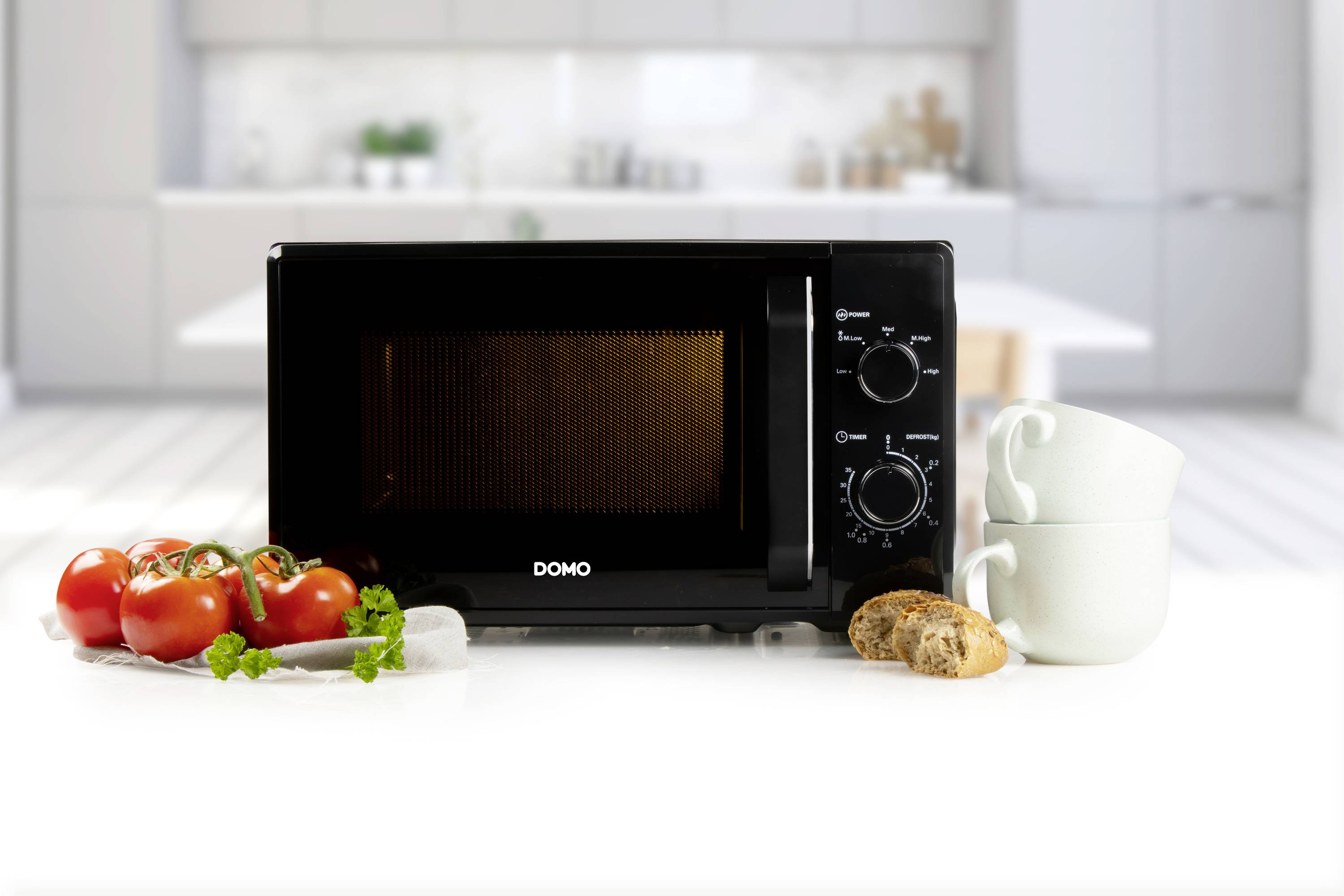 A microwave stands on a kitchen worktop. Next to it are tomatoes, herbs, mugs and slices of bread. In the background is a modern kitchen.