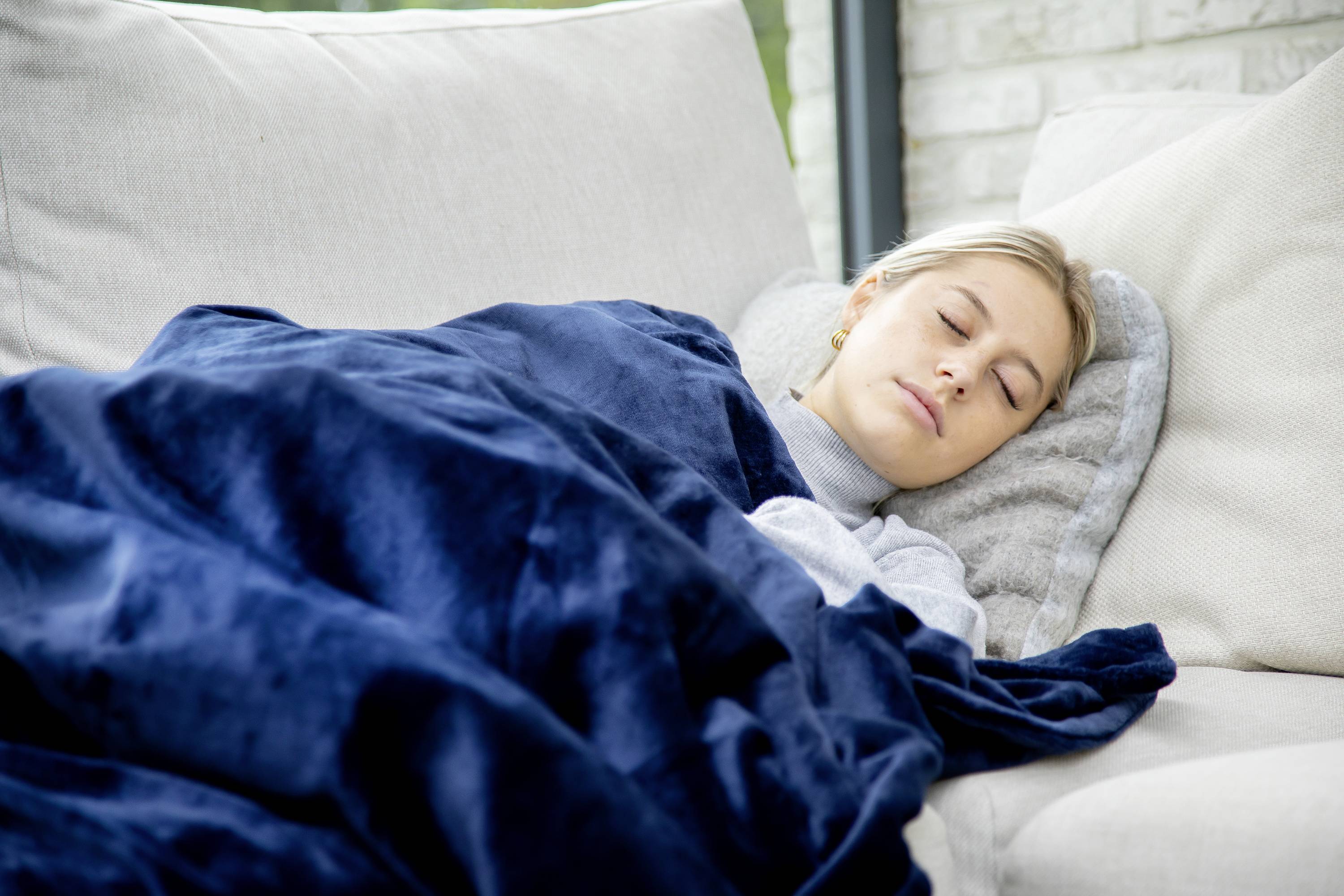A person is sleeping on a sofa, covered with a blue blanket, in a bright room with a brick wall in the background.
