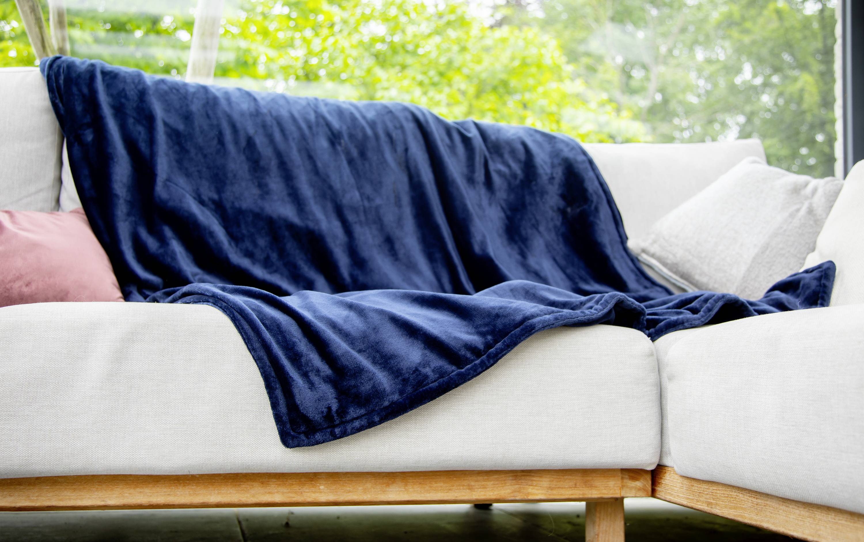 A cosy sofa with a blue throw and cushions, positioned in front of a window overlooking green trees.