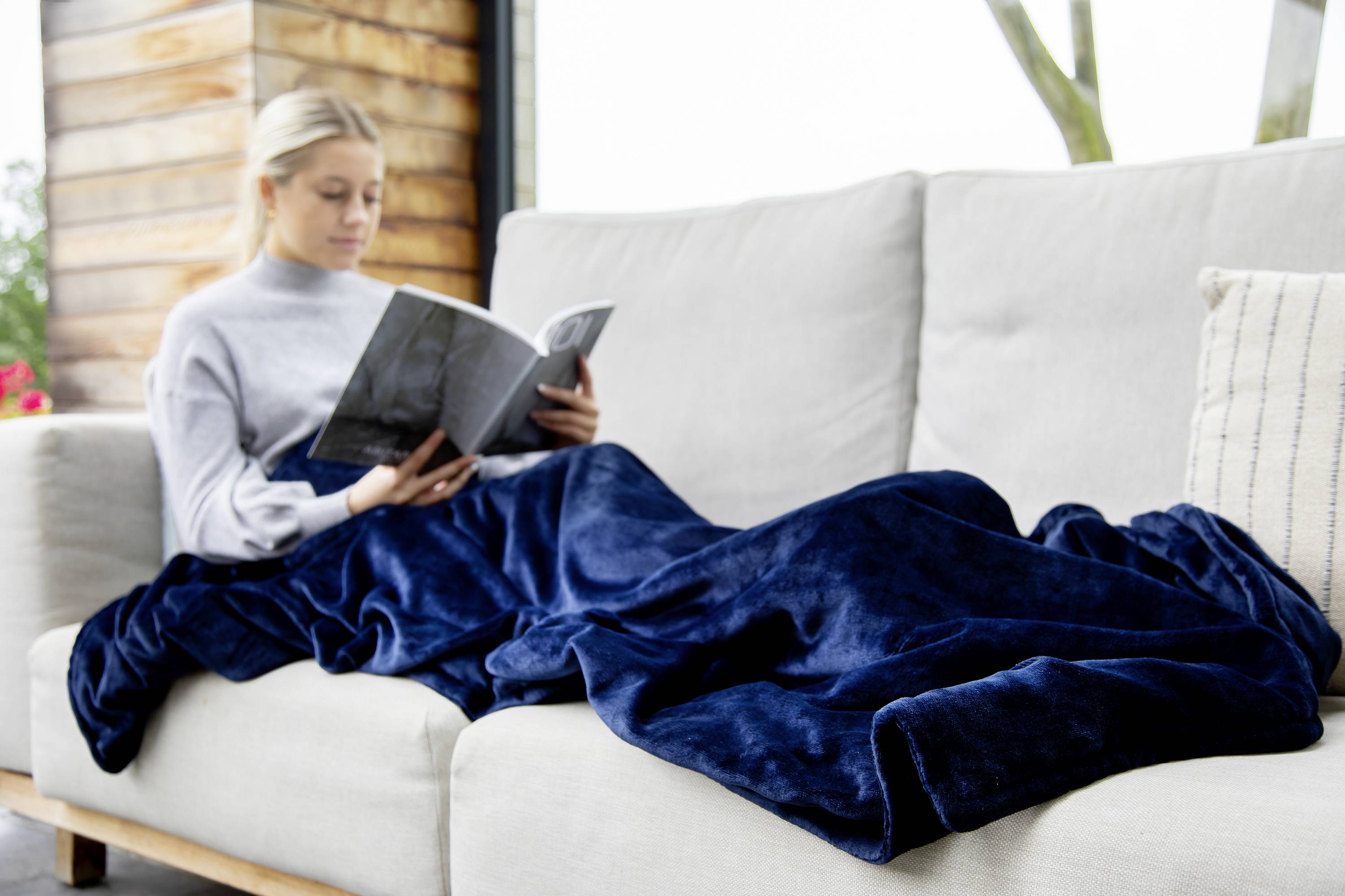 A woman is sitting on a sofa and reading a book. She is wrapped in a dark blue blanket.