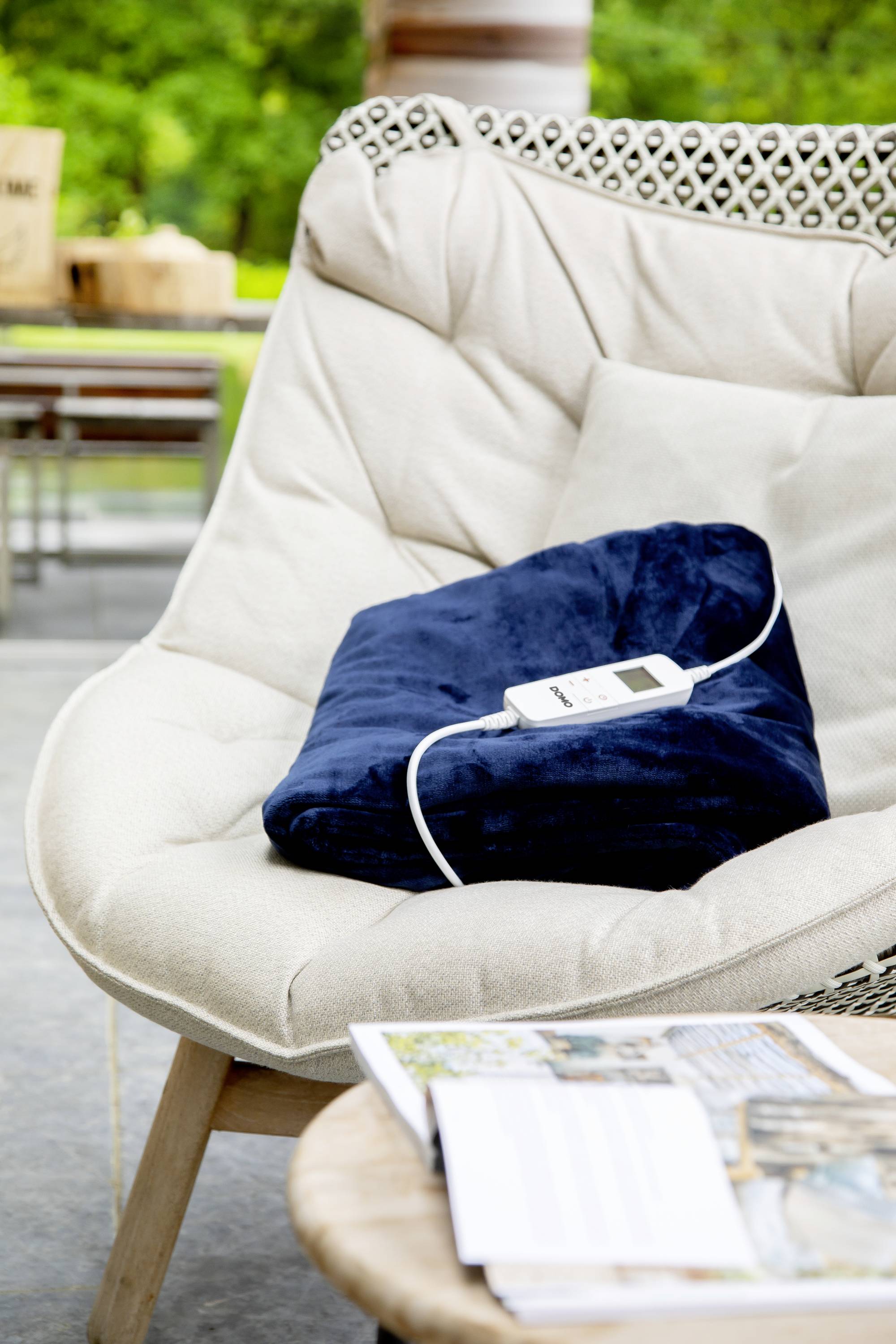 A soft, blue fabric draped on a beige armchair with a remote control resting on it, with a table and green plants in the background.