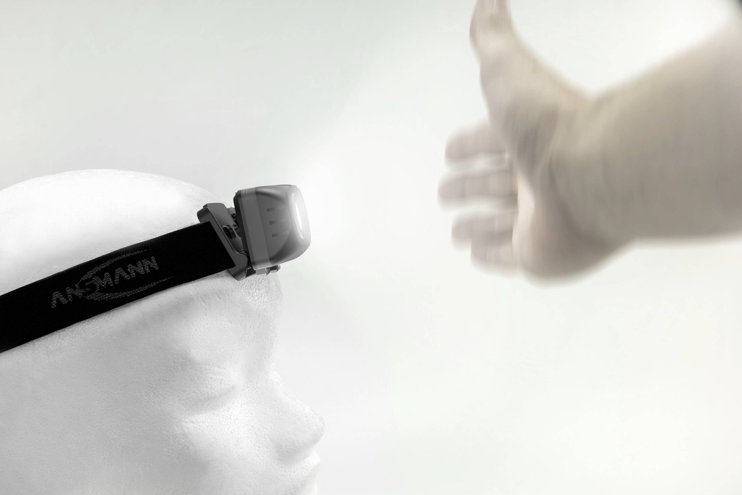 A white head with an ANSMANN head torch is illuminating an outstretched hand against a bright background.