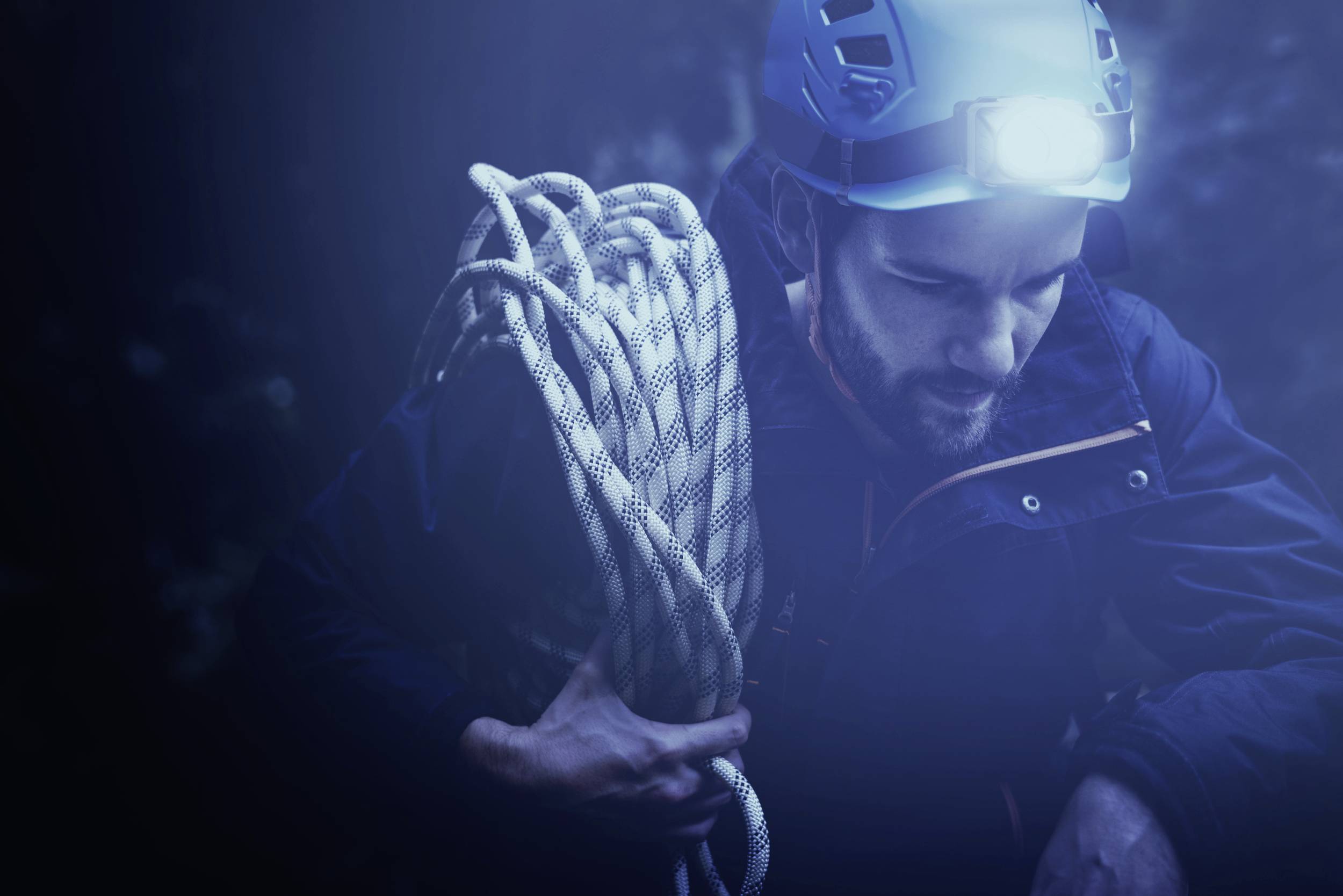 A man with climbing gear and a head torch holds a rope in a dark, unfamiliar environment.