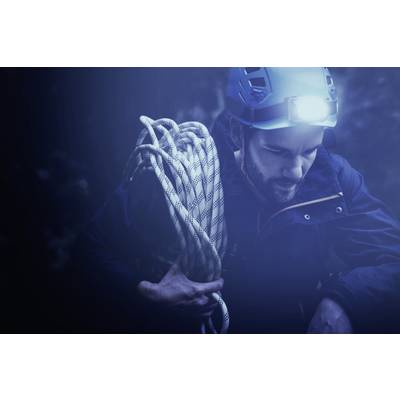 A man with climbing gear and a head torch holds a rope in a dark, unfamiliar environment.