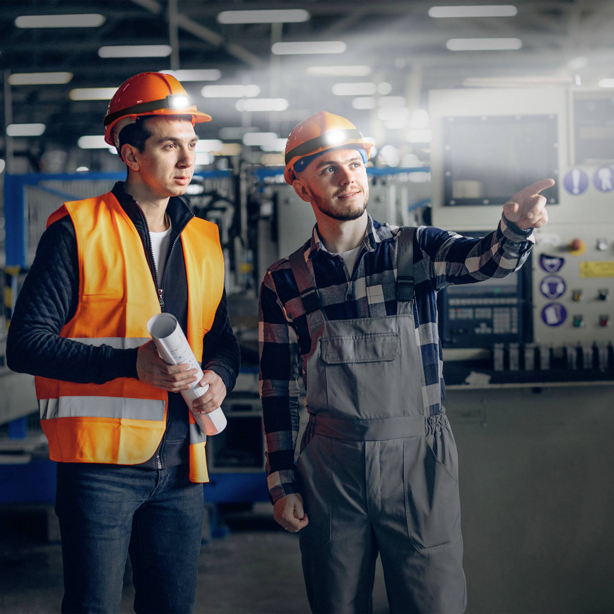 Two men in work attire and hard hats are standing in a factory hall. One is holding construction plans, while the other is pointing at machinery.