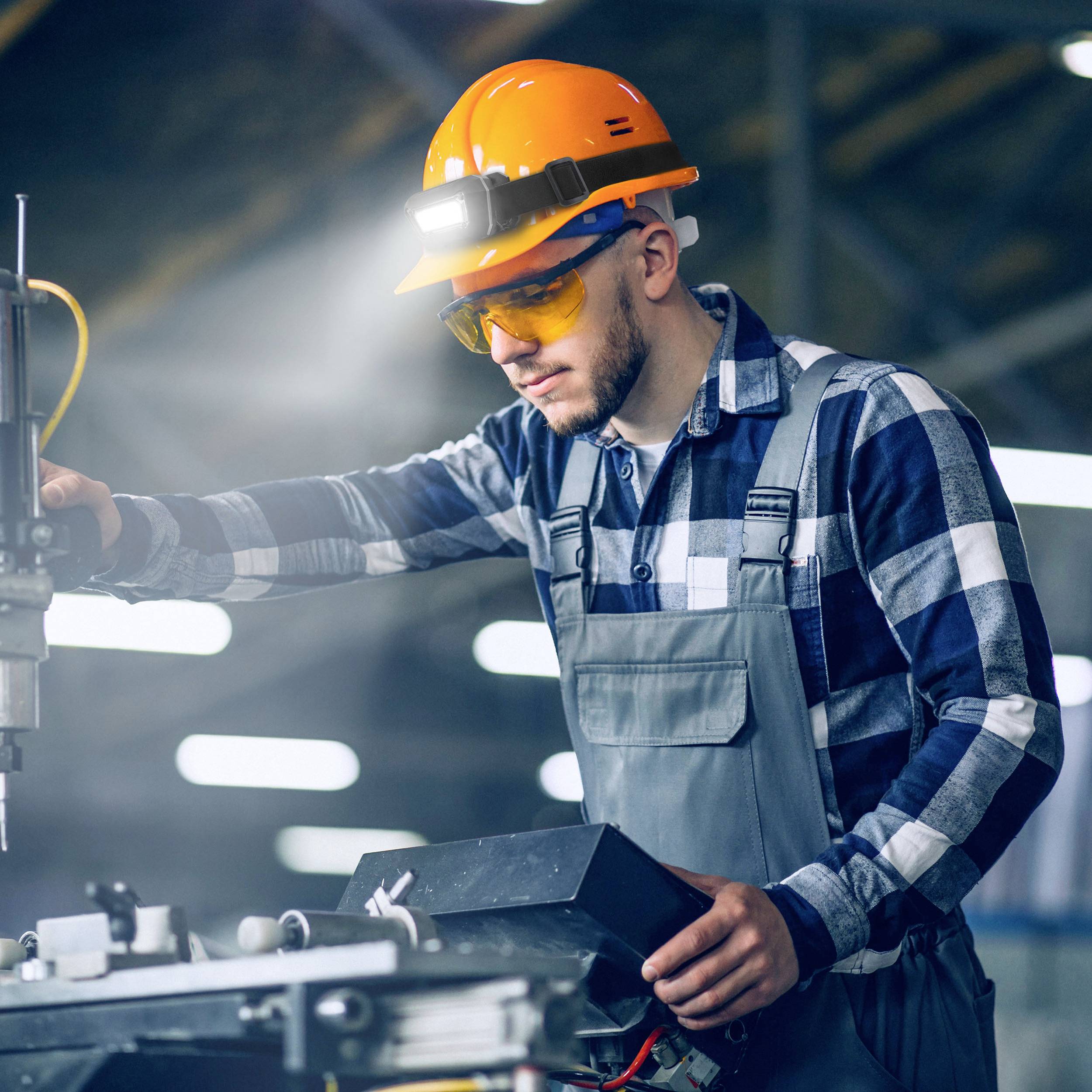 A worker wearing a hard hat and safety glasses is operating a machine in a factory. He is wearing a checked shirt and overalls.