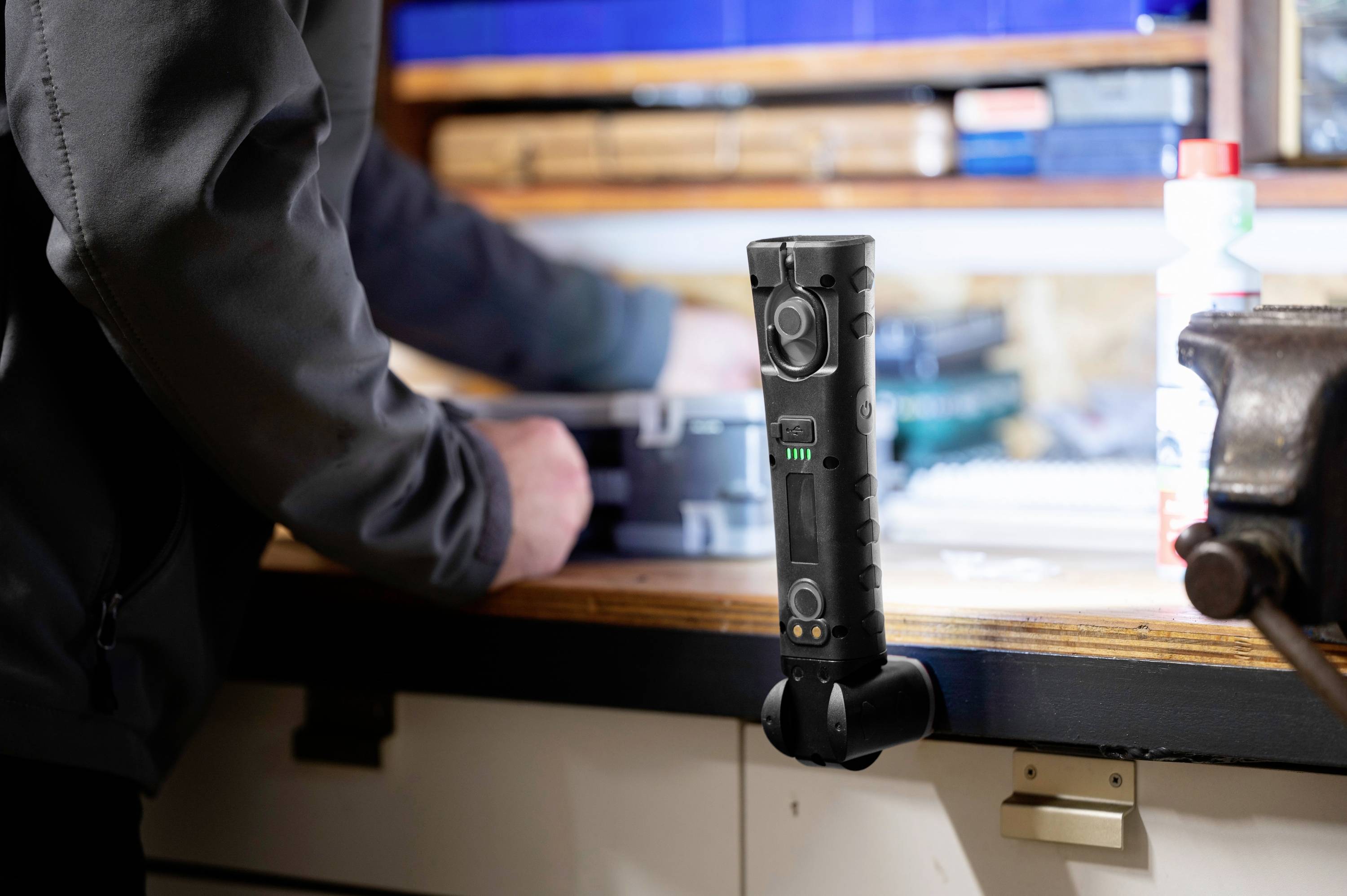 A person is standing at a workbench. In the foreground, a torch is positioned upright. Tools are blurred in the background.