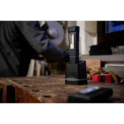 A black work lamp sits on a workbench. In the background, a blurred person can be seen working on a tool.