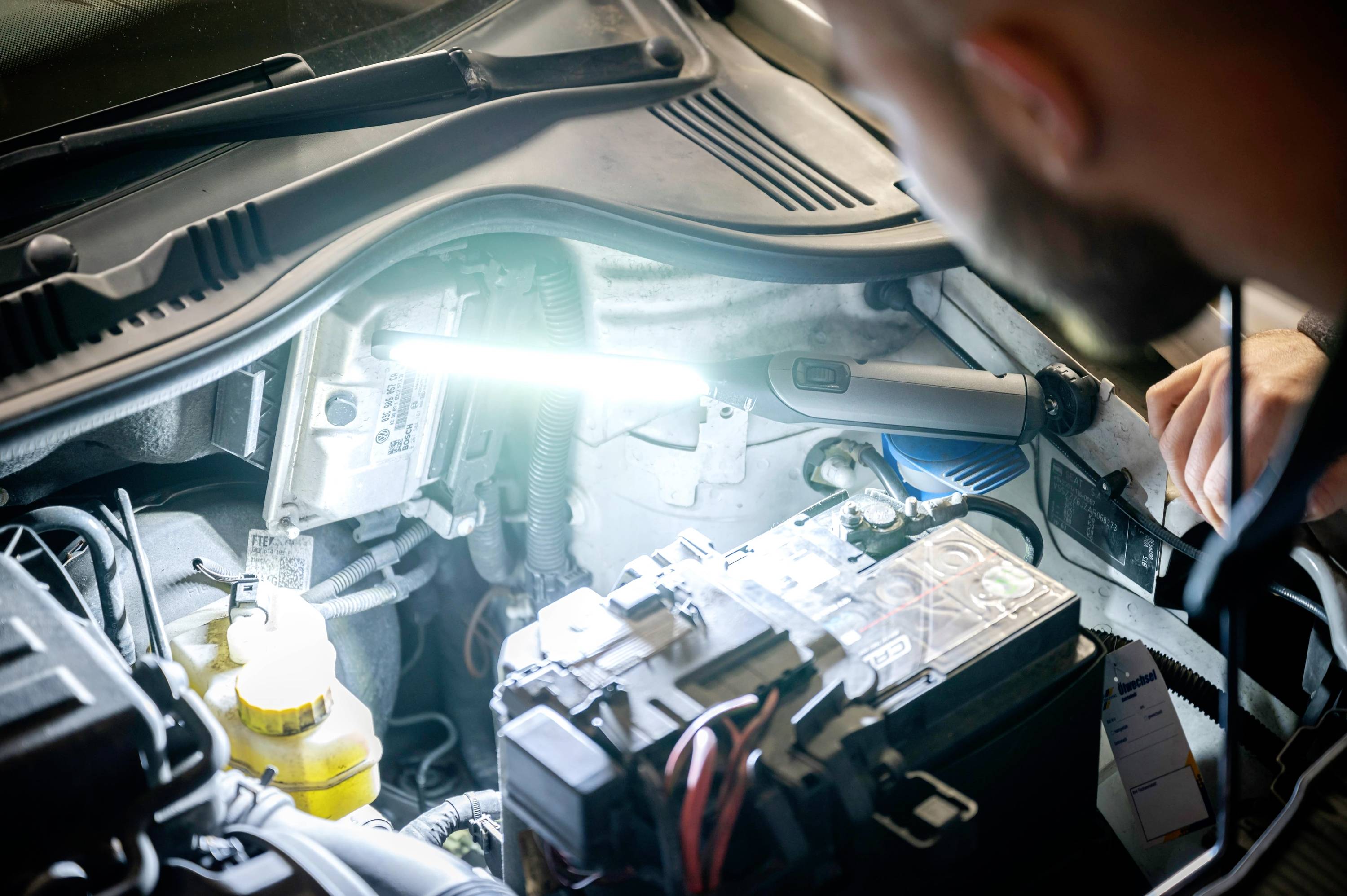 A mechanic is working under the bonnet of a car, illuminated by a lamp. Battery cables and engine components are visible.