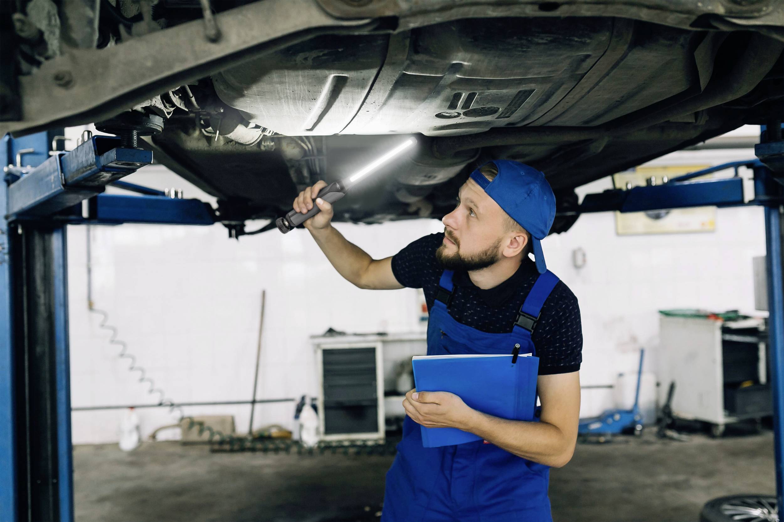 A mechanic in blue overalls is examining the underside of a car using a lamp, with the vehicle raised on a vehicle lift.