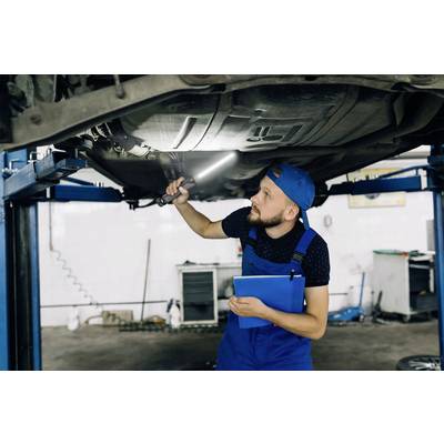 A mechanic in blue overalls is examining the underside of a car using a lamp, with the vehicle raised on a vehicle lift.