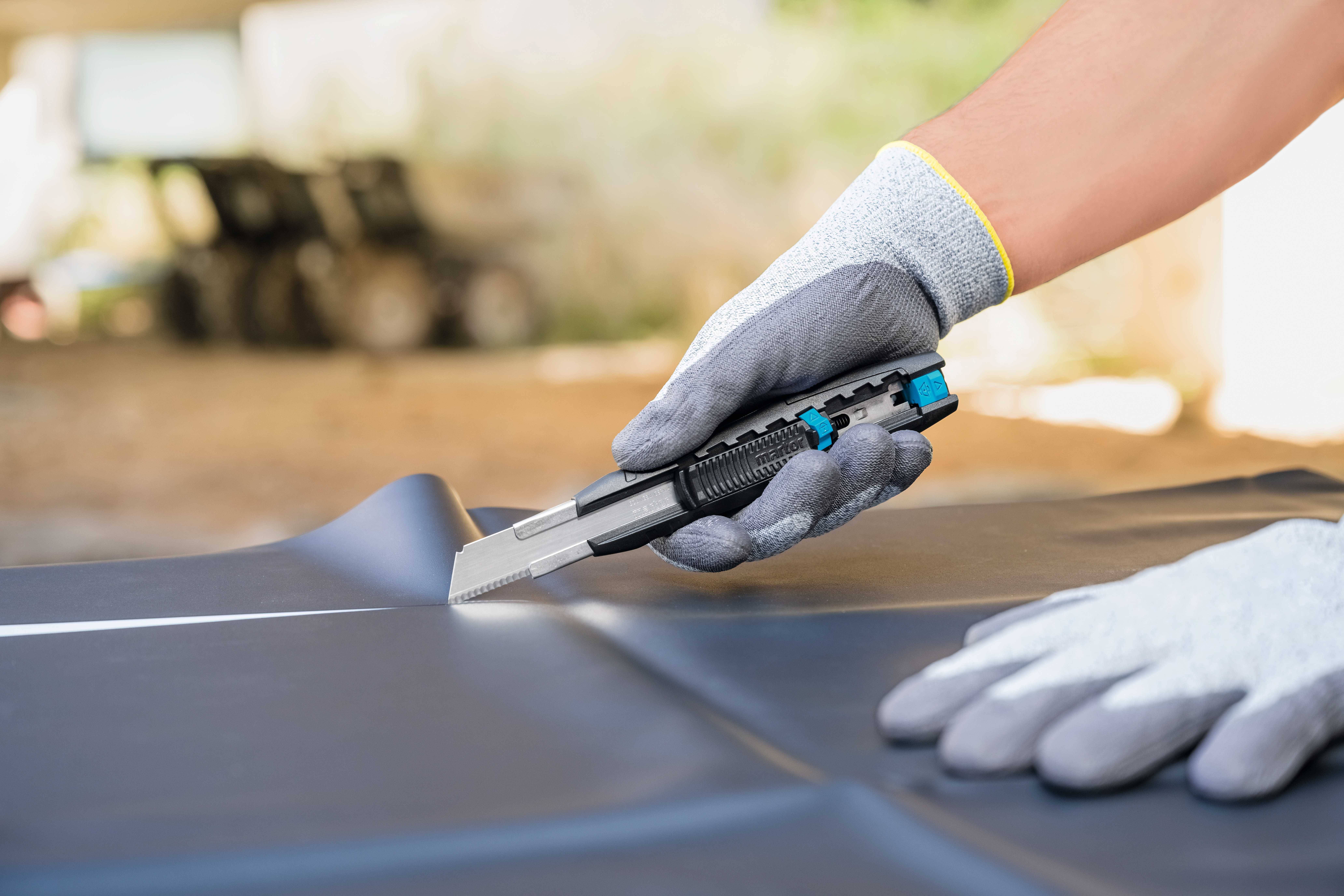 A person wearing grey gloves is cutting a black mat with a craft knife on a table. Background blurred.