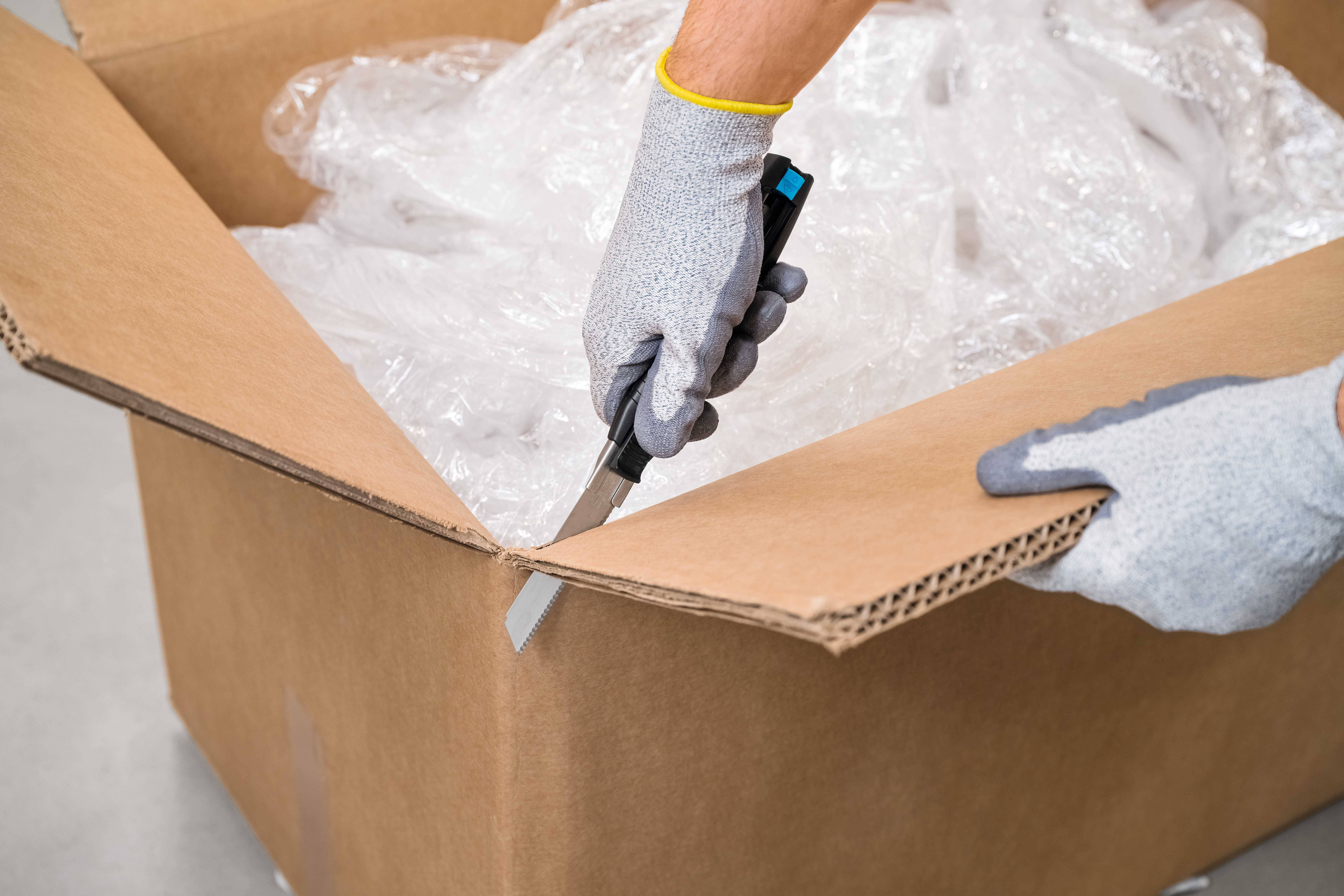 A person opens a large cardboard box with a knife, surrounded by packaging material.