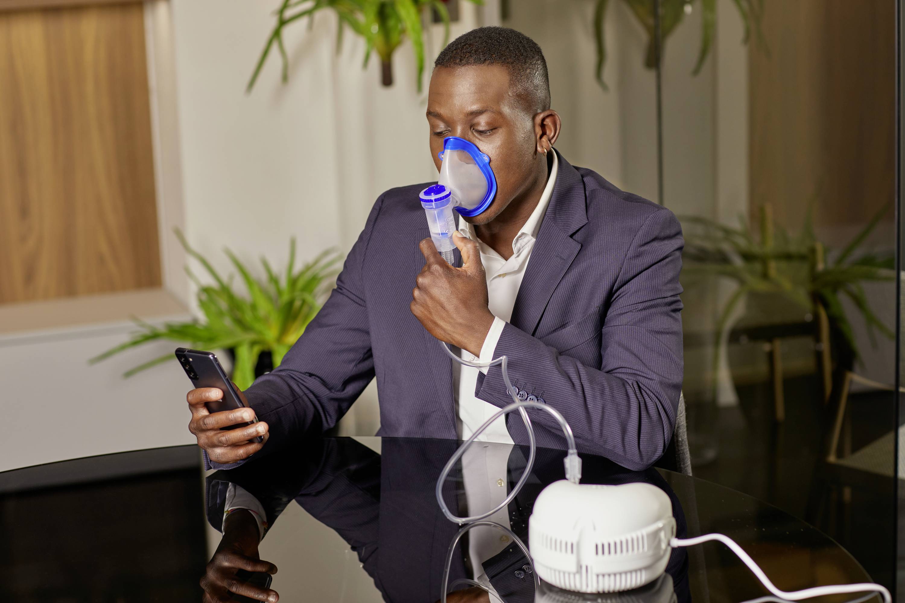 A person is sitting at a table using an inhalation device while looking at a mobile phone. Plants are visible in the background.